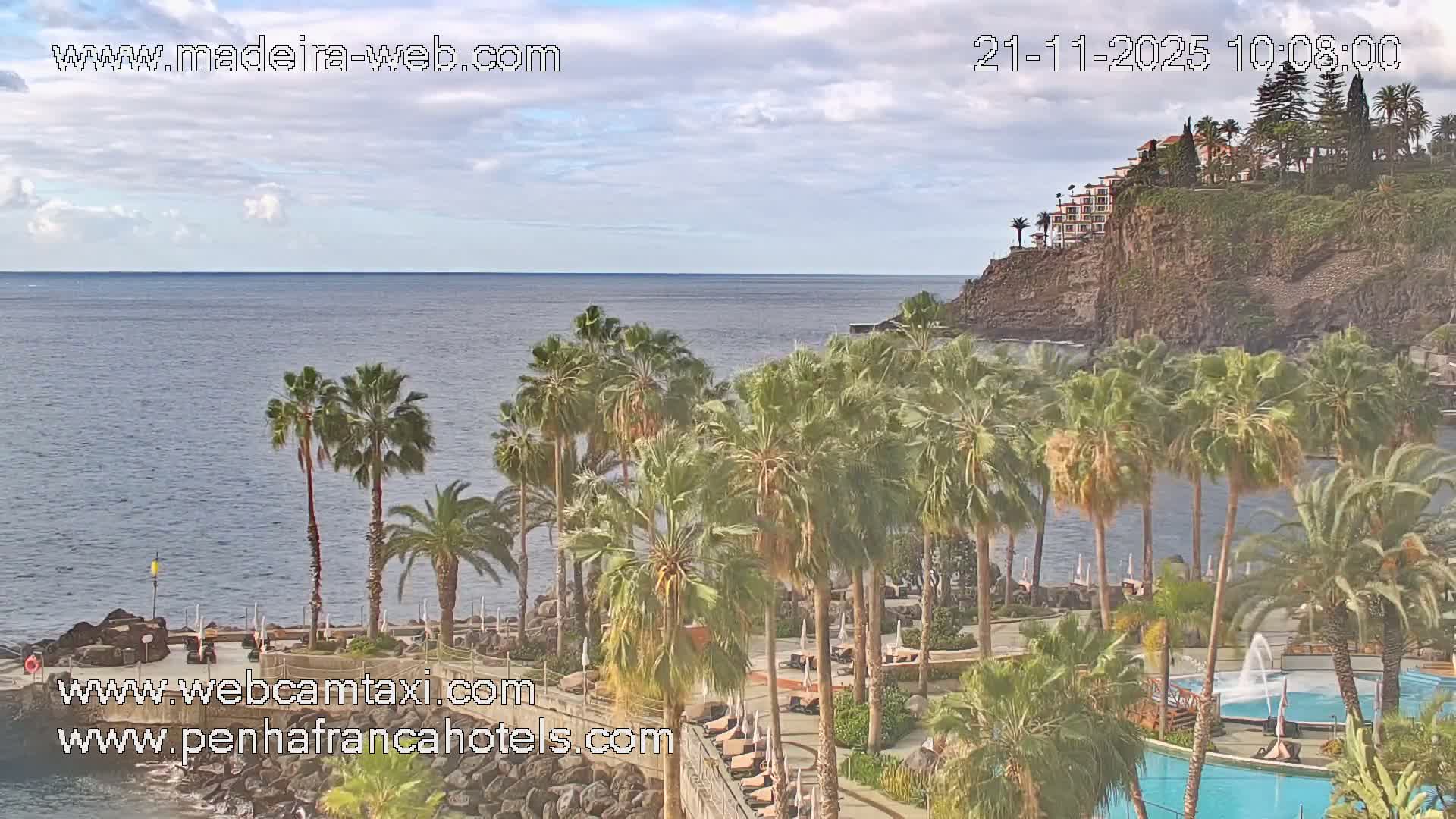 Under a partly cloudy sky, a tranquil ocean stretches beyond a rocky shore lined with numerous palm trees, a resort swimming pool, and a multi-story building perched on a distant cliff.