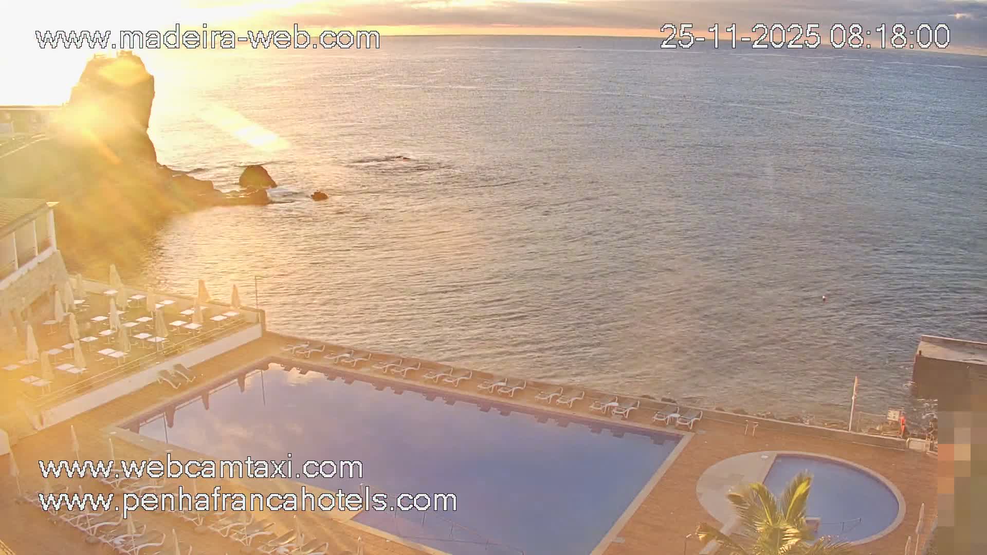 A bright, golden sunrise illuminates a coastal resort featuring large swimming pools, a lounging area with umbrellas, and a rocky coastline bordering the ocean under a partly cloudy sky.