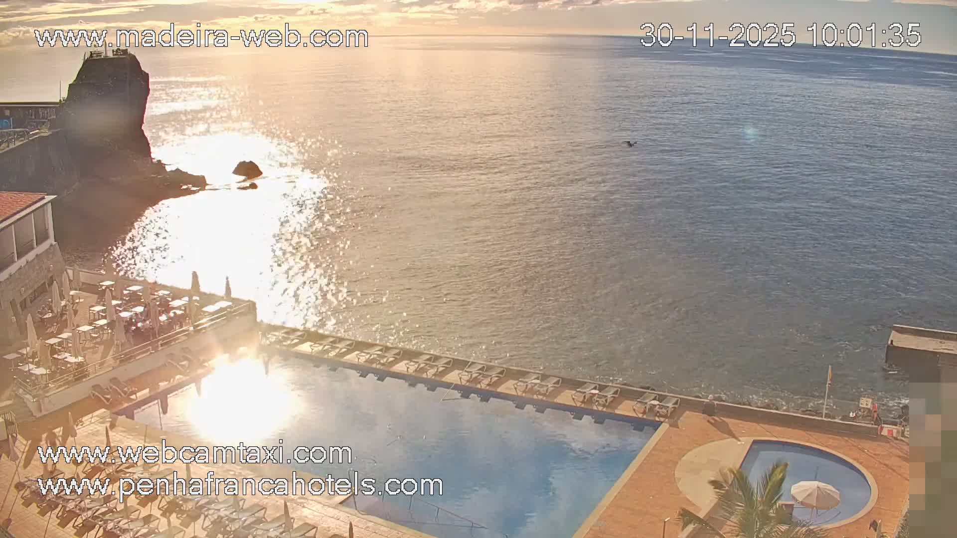 On a clear and sunny day, a coastal resort with multiple swimming pools, an outdoor restaurant, and lounge chairs overlooks a sparkling ocean featuring a prominent rock formation and a distant swimmer.
