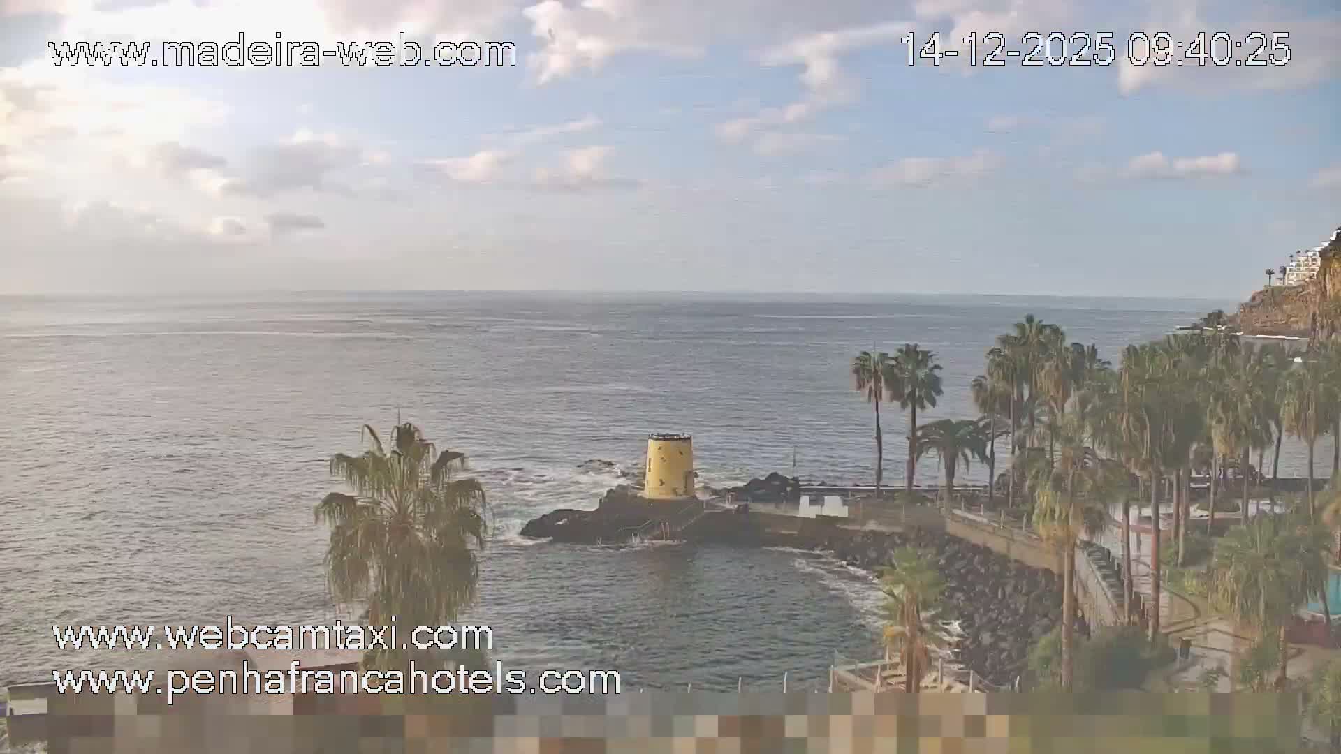 On a clear and sunny day, a coastal resort with multiple swimming pools, an outdoor restaurant, and lounge chairs overlooks a sparkling ocean featuring a prominent rock formation and a distant swimmer.