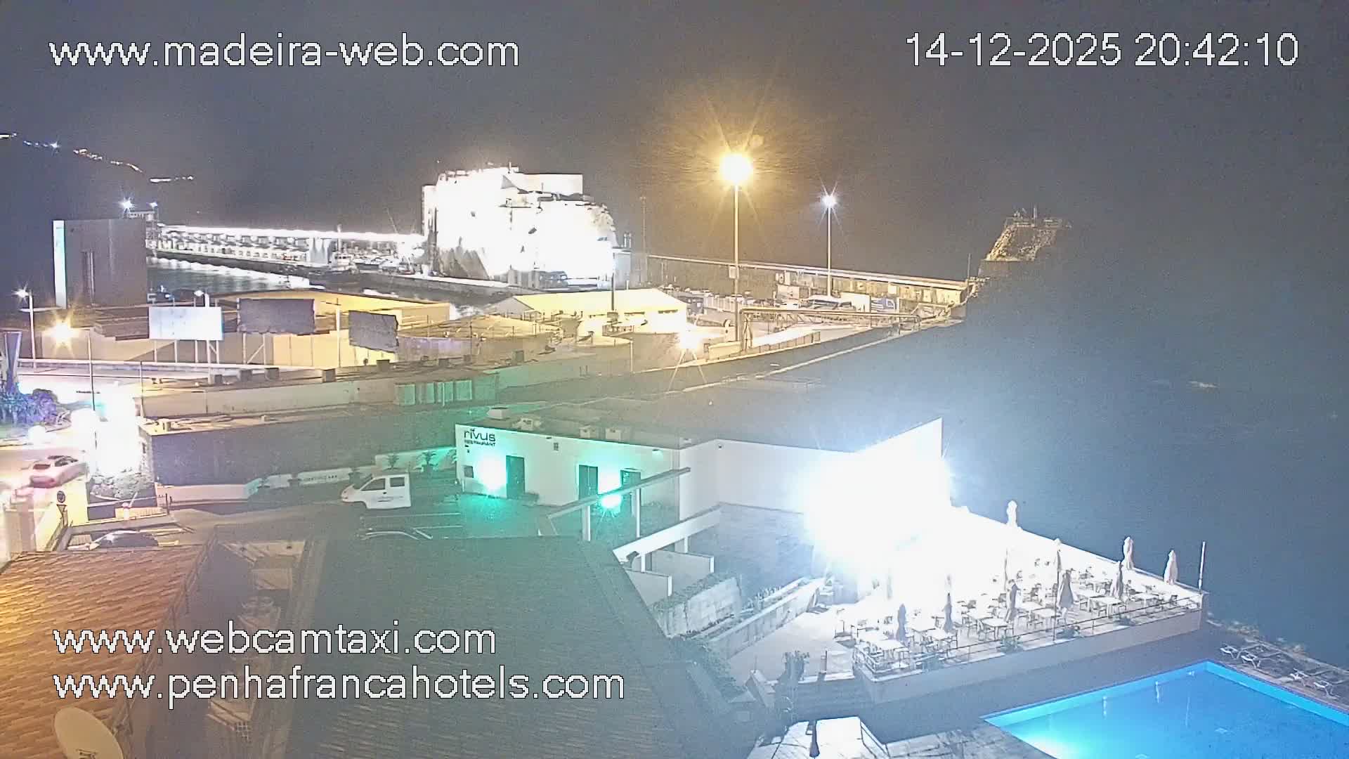 On a clear and sunny day, a coastal resort with multiple swimming pools, an outdoor restaurant, and lounge chairs overlooks a sparkling ocean featuring a prominent rock formation and a distant swimmer.