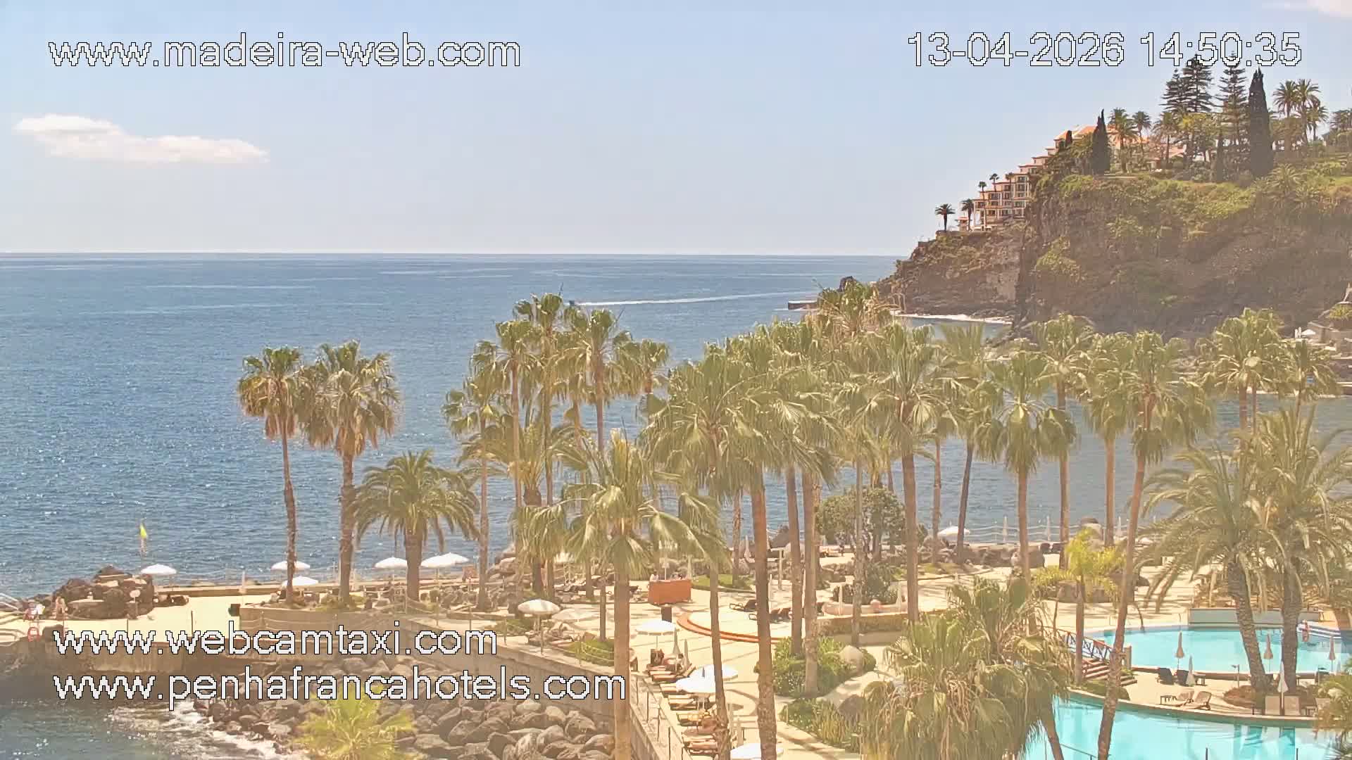On a clear and sunny day, a coastal resort with multiple swimming pools, an outdoor restaurant, and lounge chairs overlooks a sparkling ocean featuring a prominent rock formation and a distant swimmer.