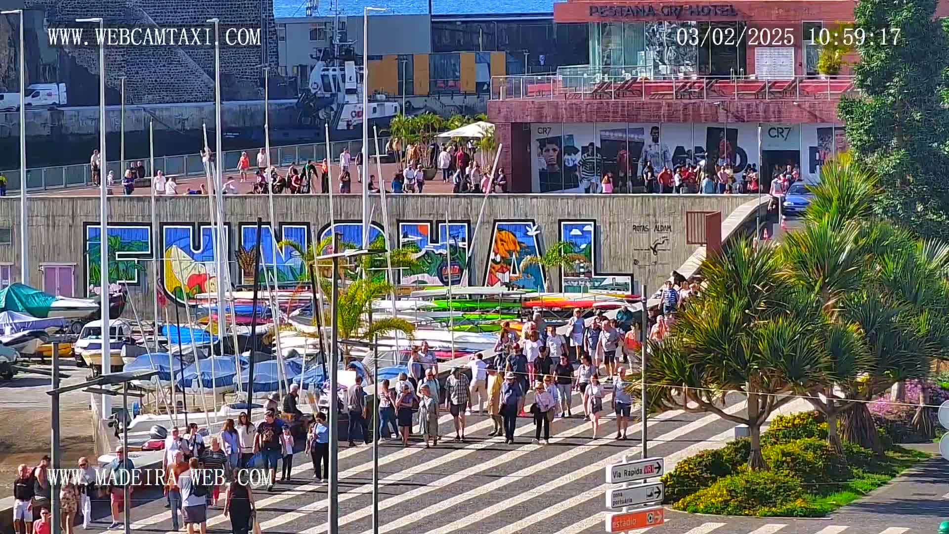 A sunny harbor scene shows a crowd of people walking on a pedestrian crossing in front of a large graffiti mural that says "Funchal," with numerous boats and yachts docked behind them.