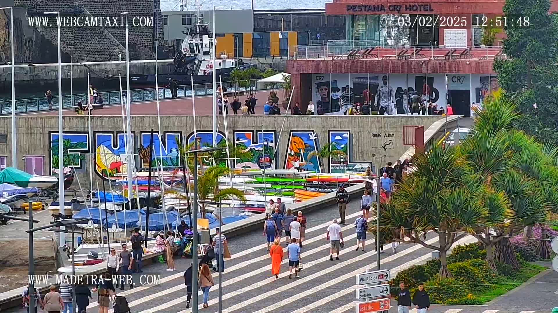 A waterfront area with a large mural, numerous small boats docked, and pedestrians walking on a crosswalk under a sunny sky.