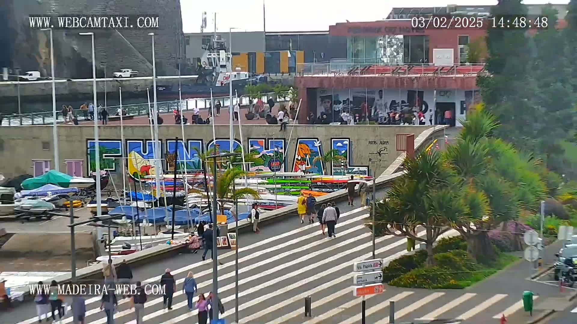 A harbor scene with numerous boats docked along a waterfront, featuring a large mural and pedestrians walking on a crosswalk, under a partly sunny sky.