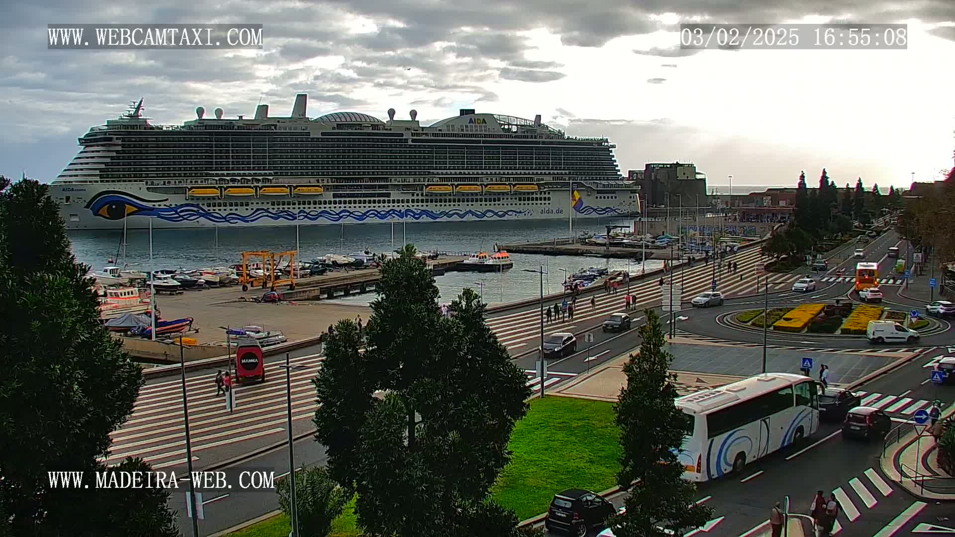 A large cruise ship is docked at a port, with overcast skies and several vehicles and pedestrians visible on the streets nearby.