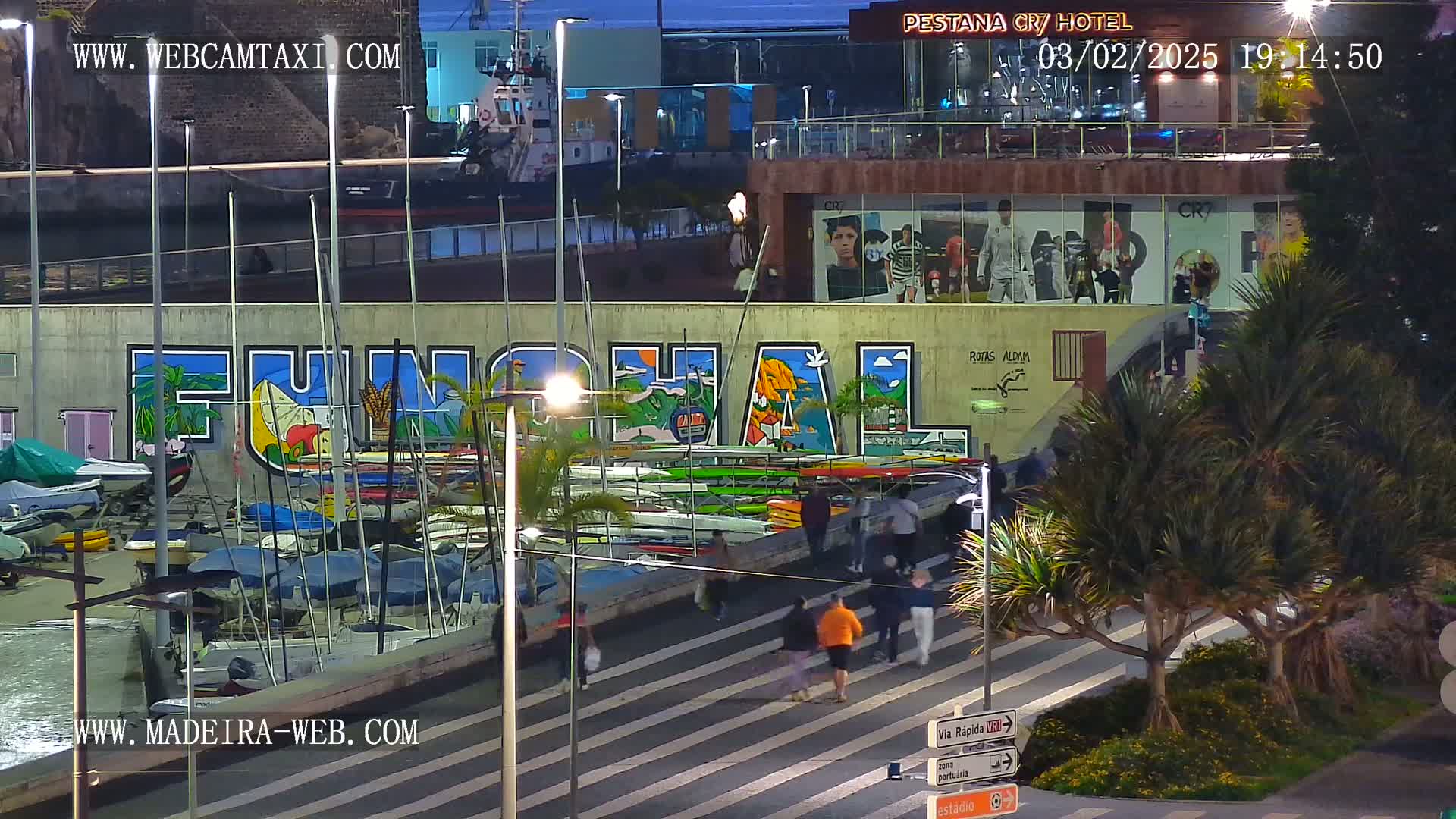 A nighttime harbor scene shows a large mural of the word "FUNCHAL" on a wall overlooking numerous small sailboats, with pedestrians walking along a crosswalk nearby under streetlights.