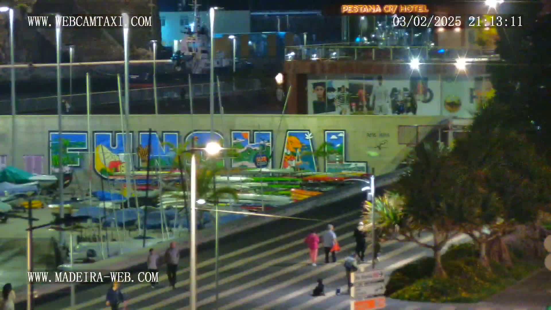 A nighttime view of a harbor with numerous boats and kayaks, a large mural depicting the word "Funchal," and pedestrians walking along a walkway beside a building.