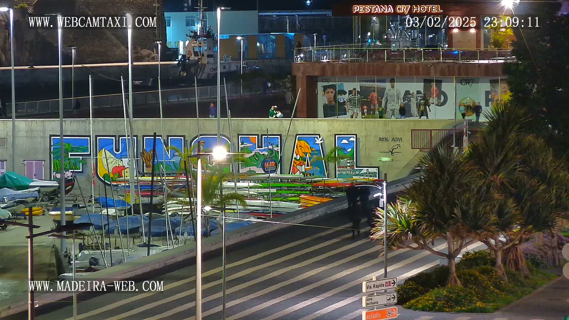 A nighttime view of a marina with numerous small boats, a large mural depicting the word "Funchal," and palm trees, under clear skies.