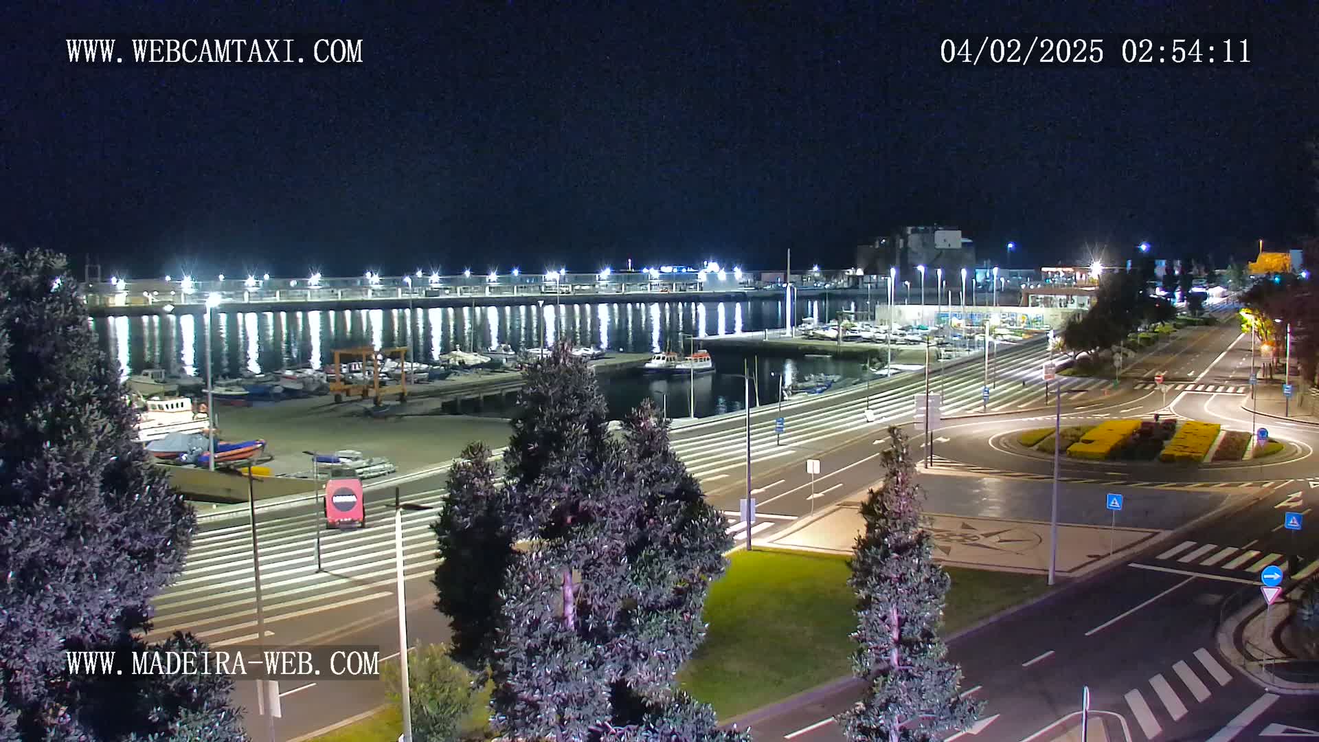 A nighttime view of a harbor filled with boats, adjacent to a roadway with a roundabout, under a clear sky.