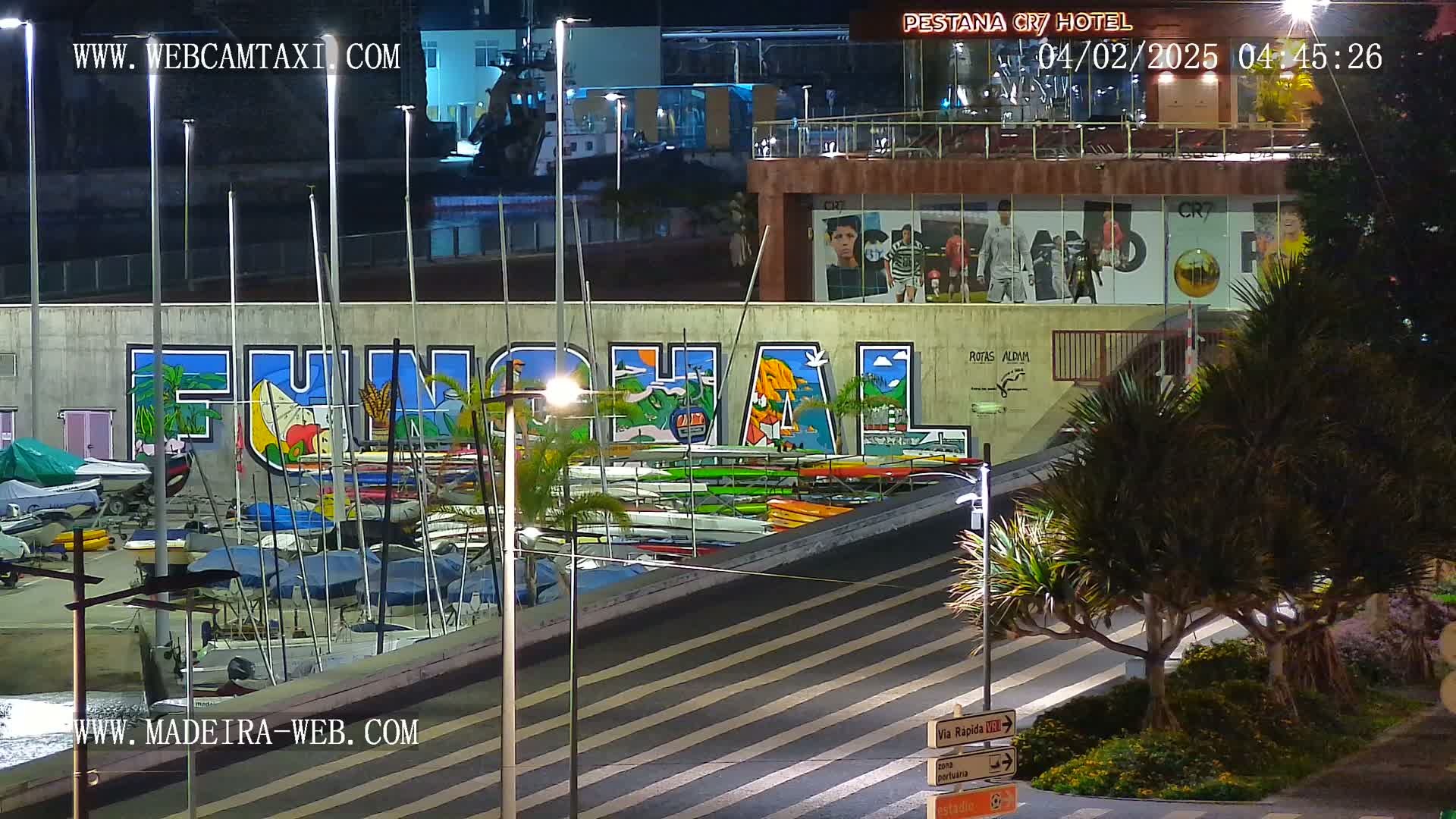 A nighttime view of a marina with numerous sailboats and kayaks, a large mural depicting the word "FUNCHAL," and a building with illuminated signage.
