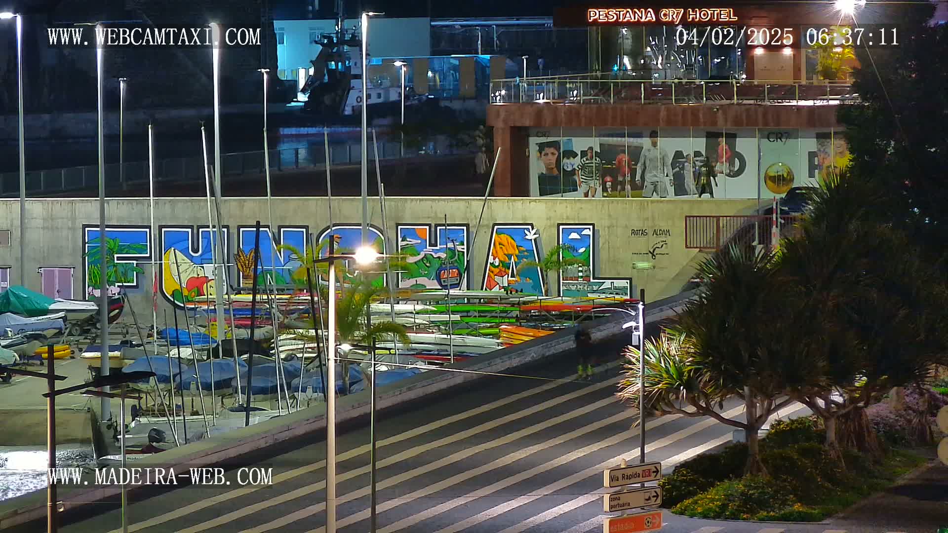 A nighttime view of a marina with numerous boats and kayaks, a large mural depicting the word "FUNCHAL," and palm trees, under artificial light.