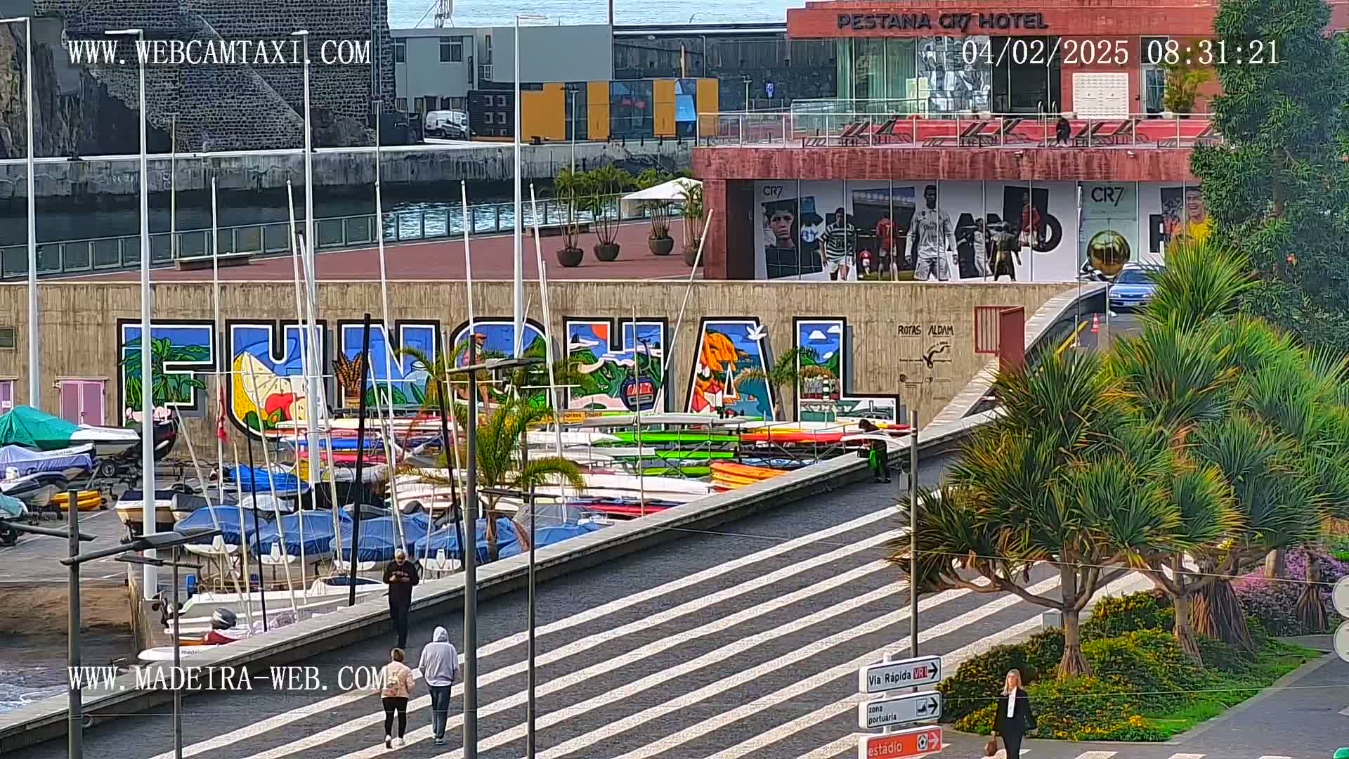 A sunny harbor scene shows numerous sailboats and kayaks docked along a wall painted with a large mural of the word "Funchal," with several people walking nearby.