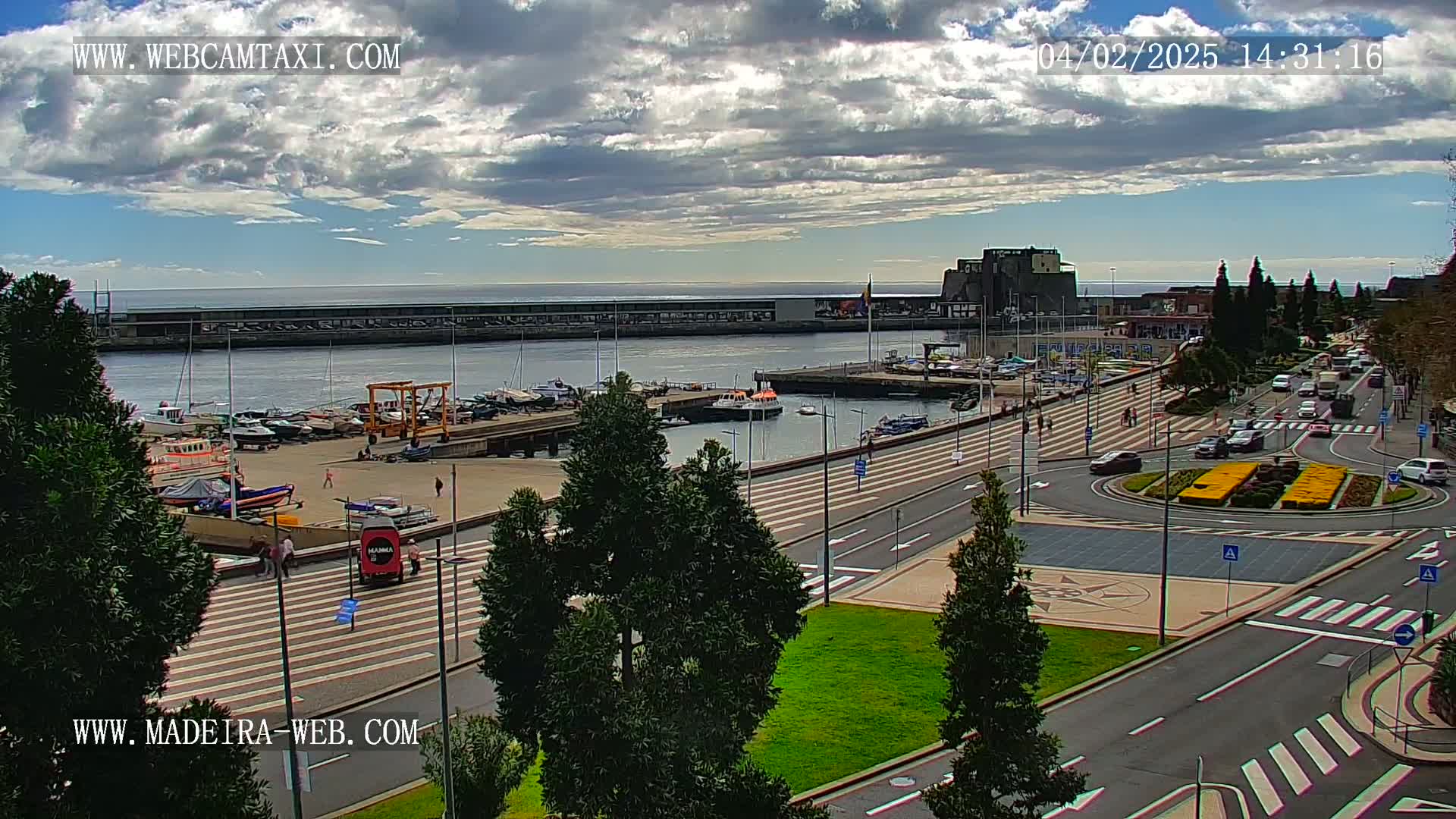A harbor with many boats, a paved area with cars and pedestrians, and green spaces, all under a partly cloudy sky.