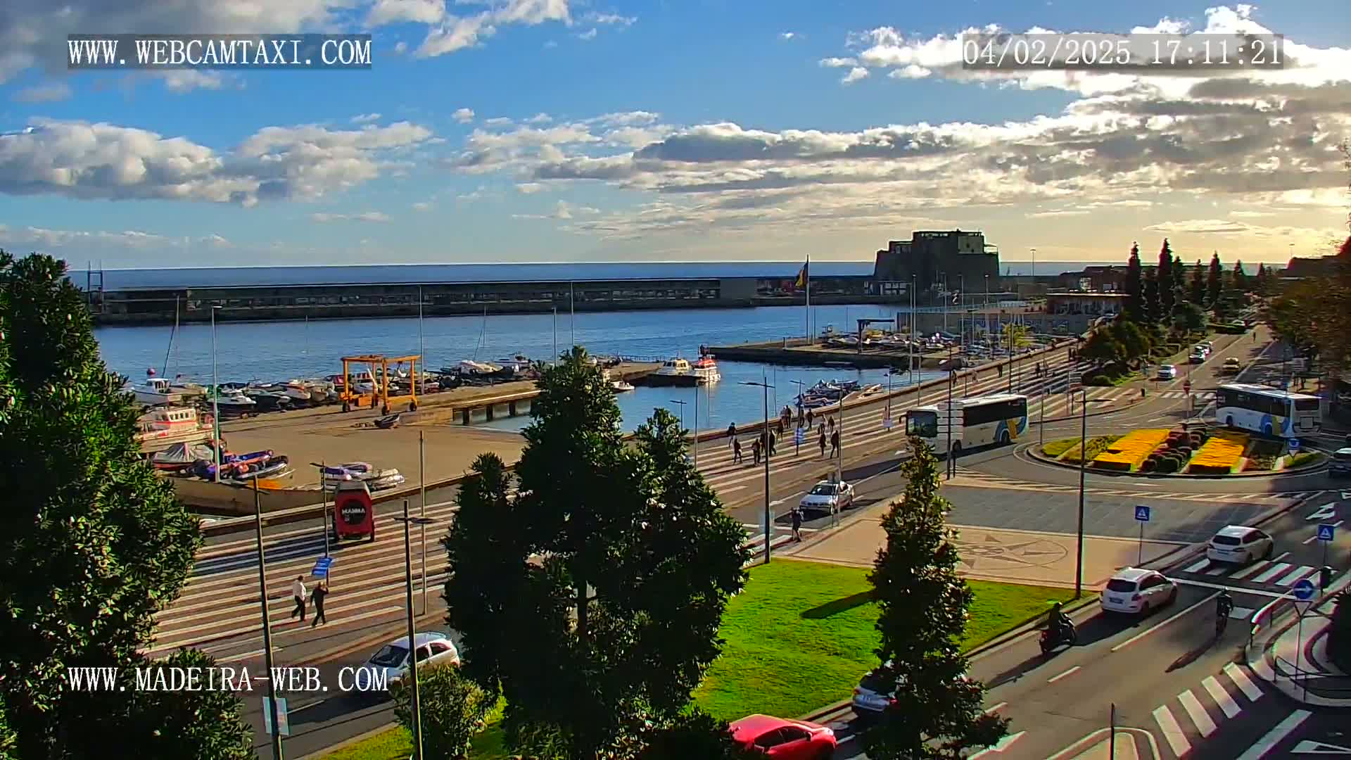 A harbor with numerous boats, a roadway with cars and buses, and pedestrians walking along the waterfront under a partly cloudy sky.