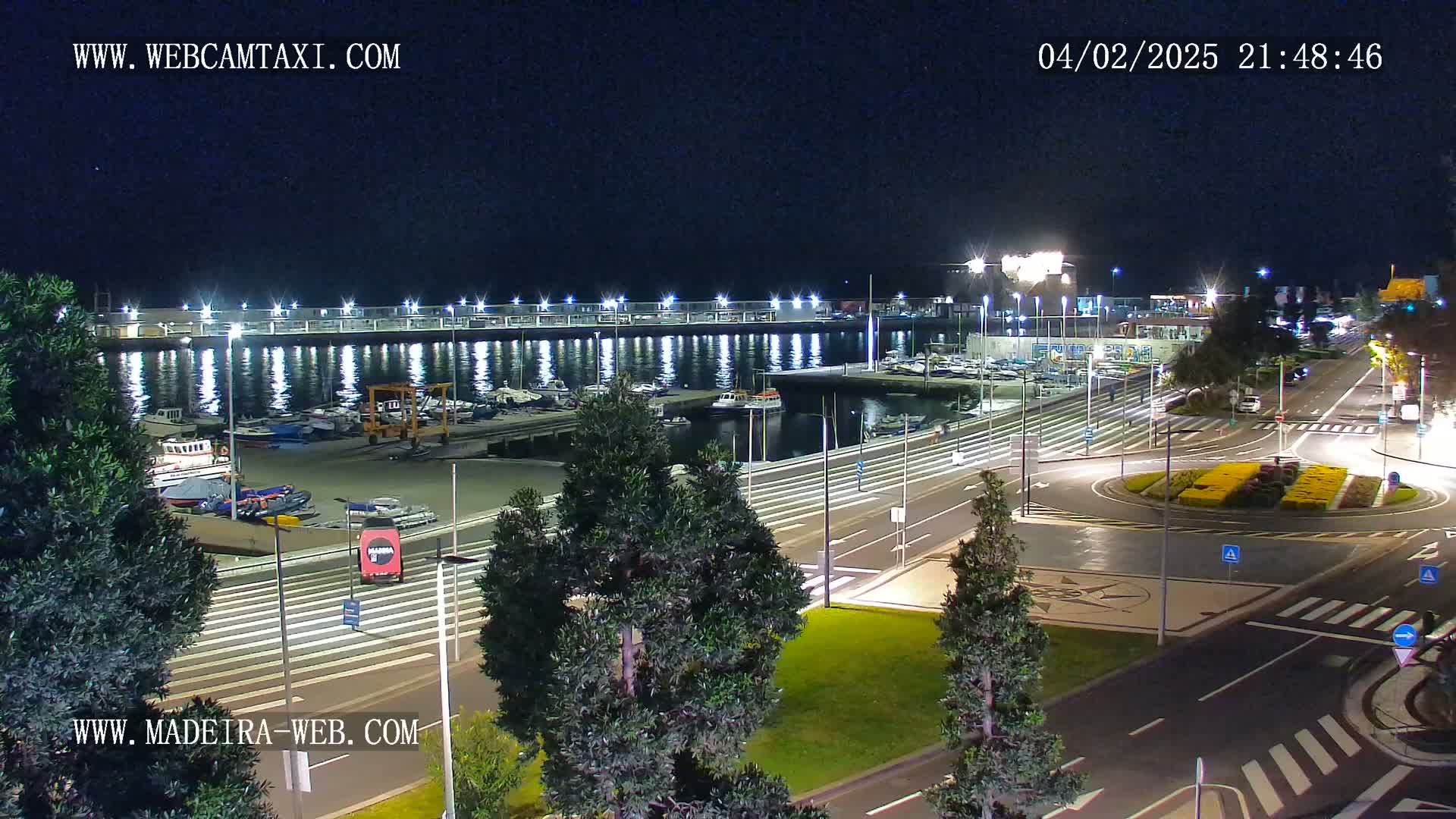 A nighttime view of a marina with boats docked, a roadway with a roundabout, and surrounding green spaces under clear skies.
