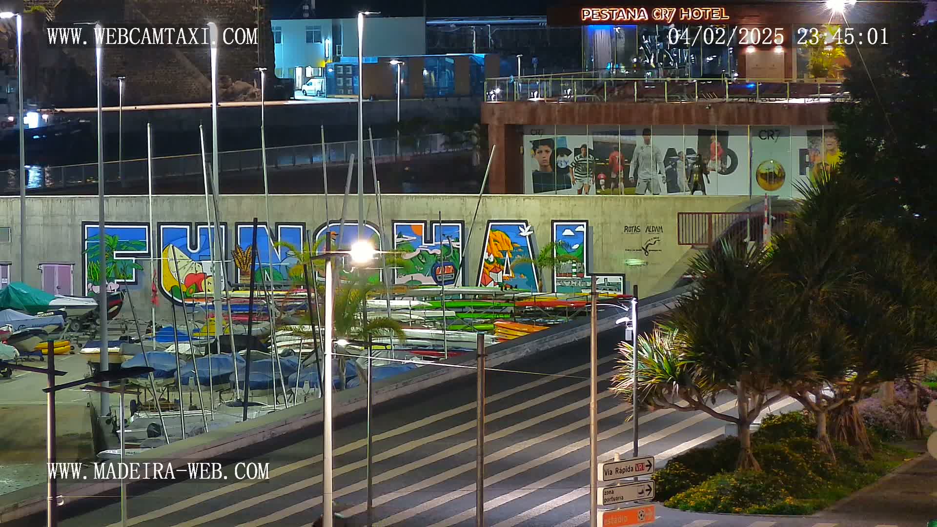 At night, a marina with numerous boats is shown beneath a large mural that says "FUNCHAL," next to a building with large windows displaying images of soccer players.
