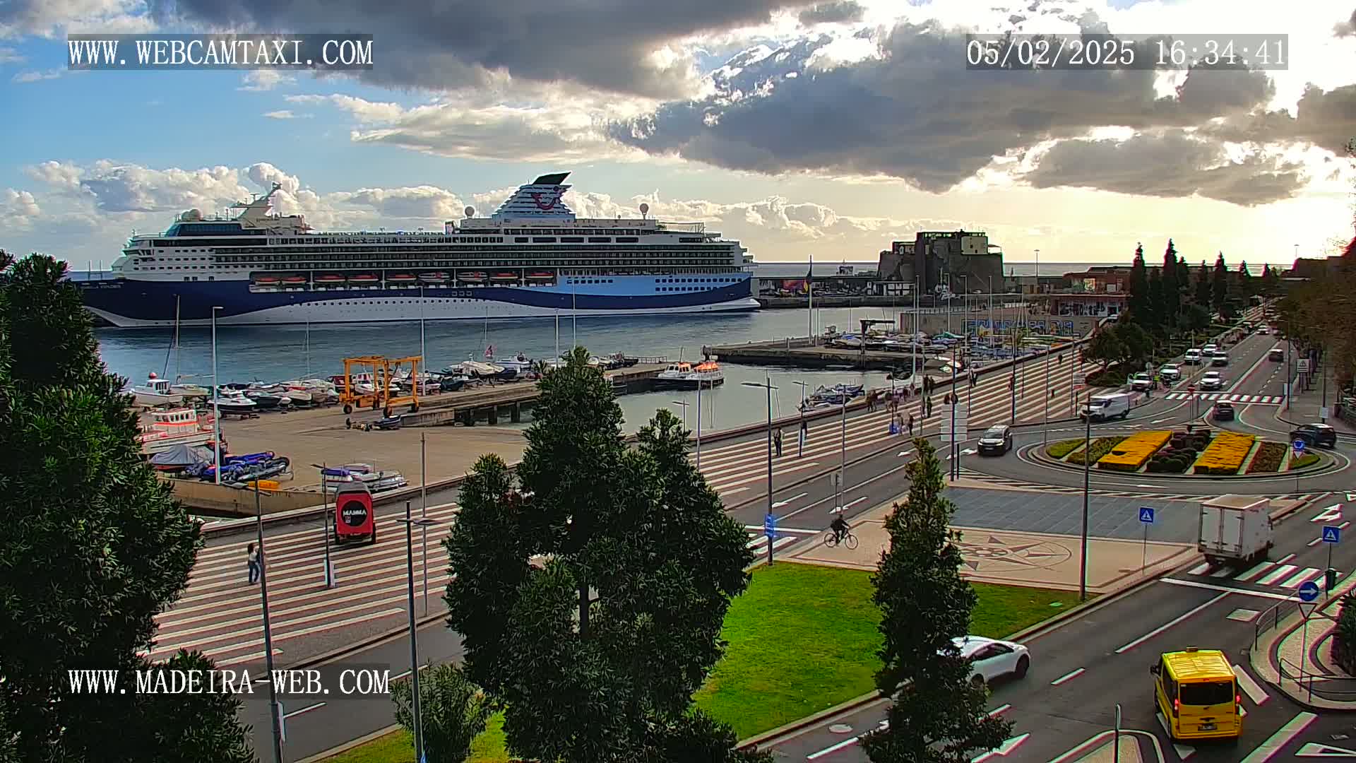 A large cruise ship is docked at a harbor, with partly cloudy skies and light traffic on the streets nearby.