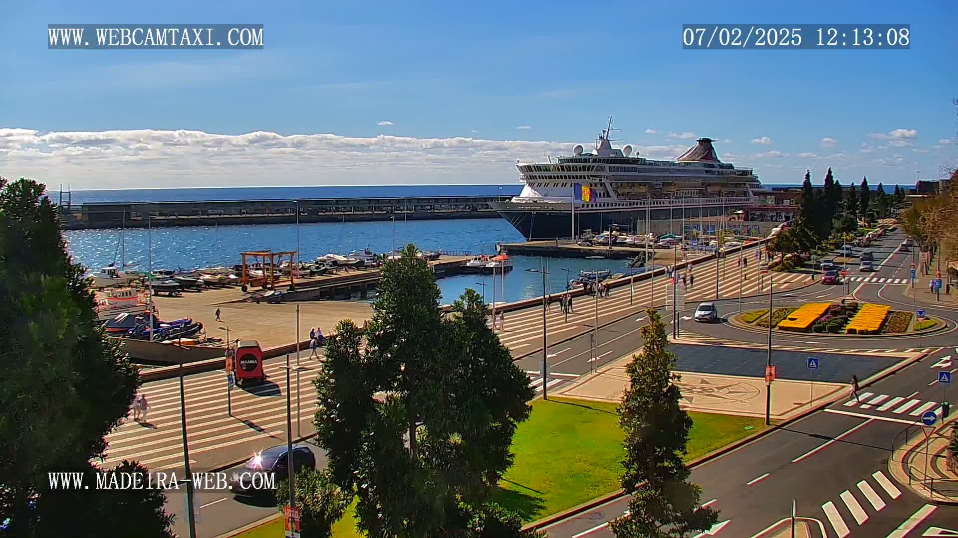 A large cruise ship is docked at a harbor on a sunny day, with pedestrians and vehicles visible on the adjacent streets and promenade.