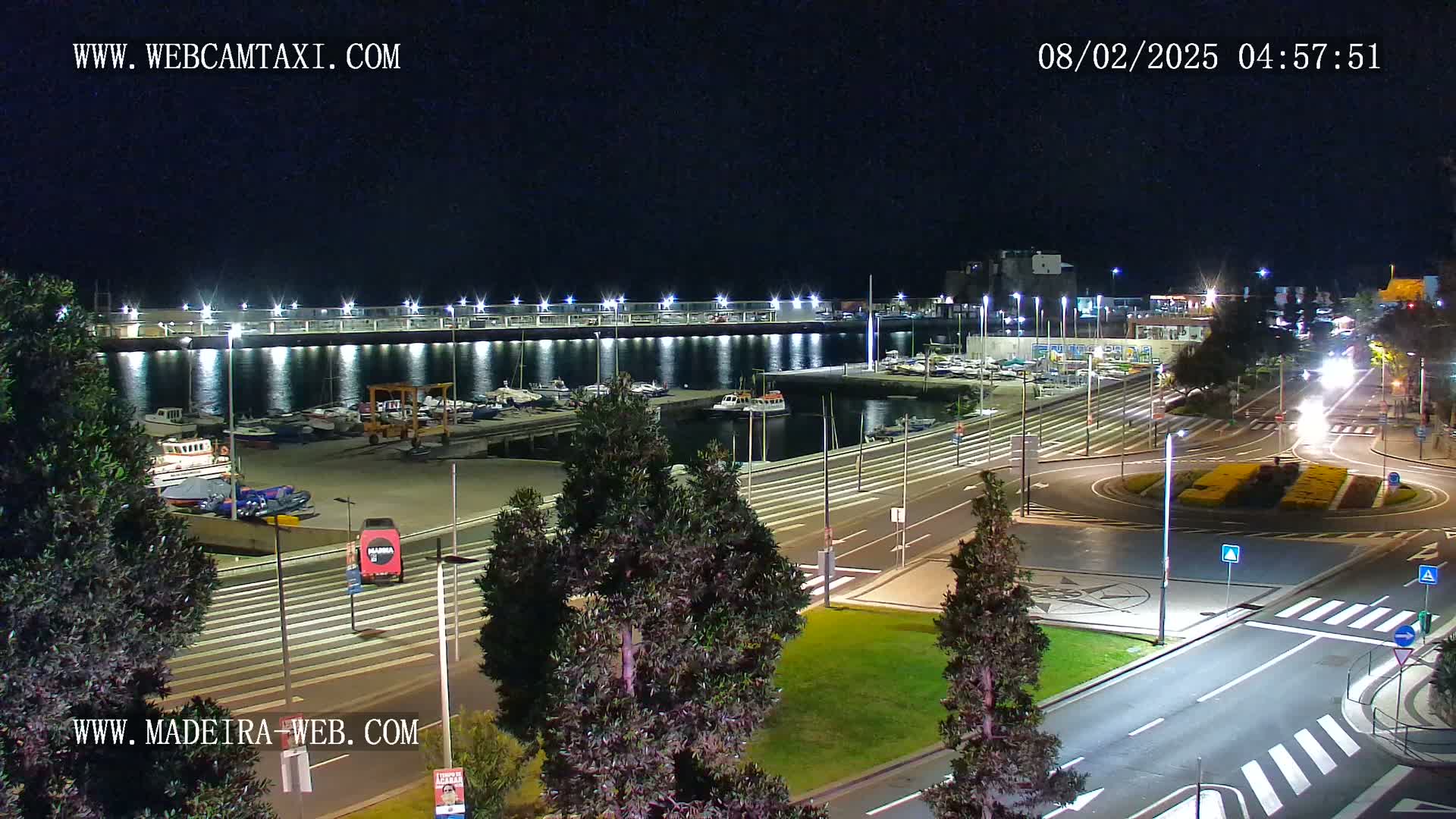 A nighttime view of a harbor filled with boats, adjacent to a roadway with a roundabout, all brightly lit.