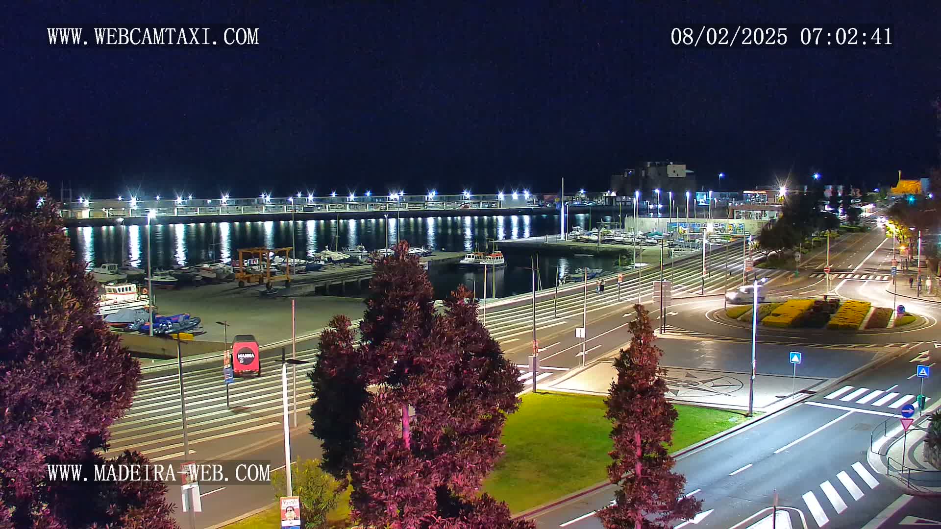 A nighttime view of a harbor filled with boats, next to a roadway with a roundabout, under a clear, dark sky.