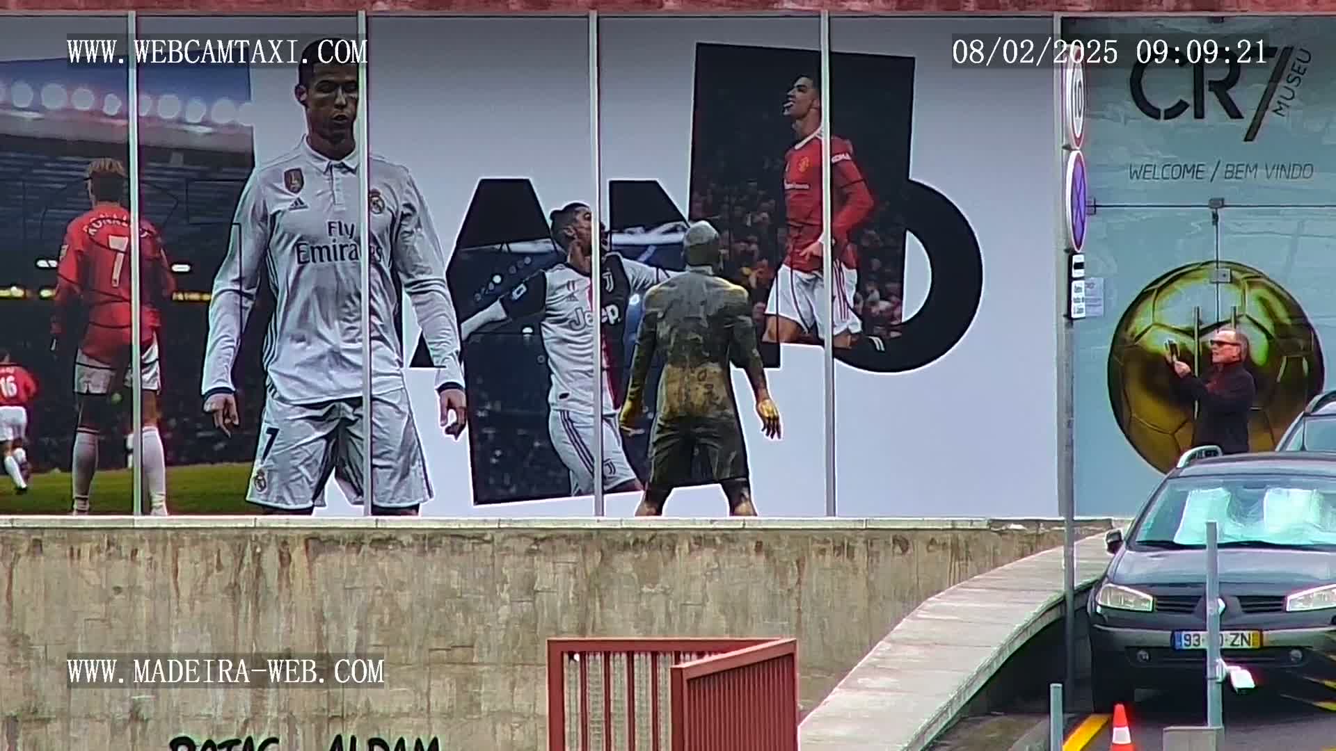A large building displays a mural of various soccer players, and a person takes a picture of a golden soccer ball near a parked car.