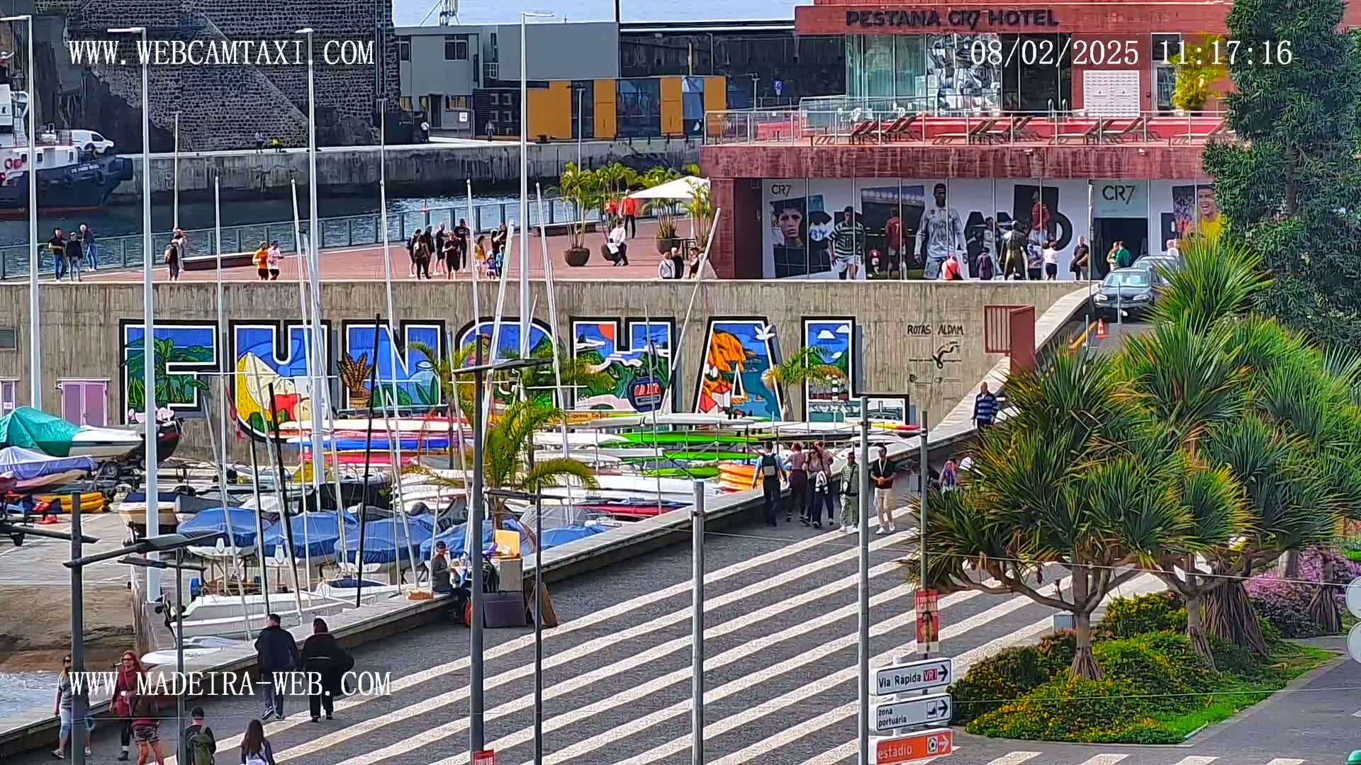 A sunny day shows a harbor filled with sailboats and kayaks next to a wall with a large mural of the word Funchal, with people walking along a paved area.