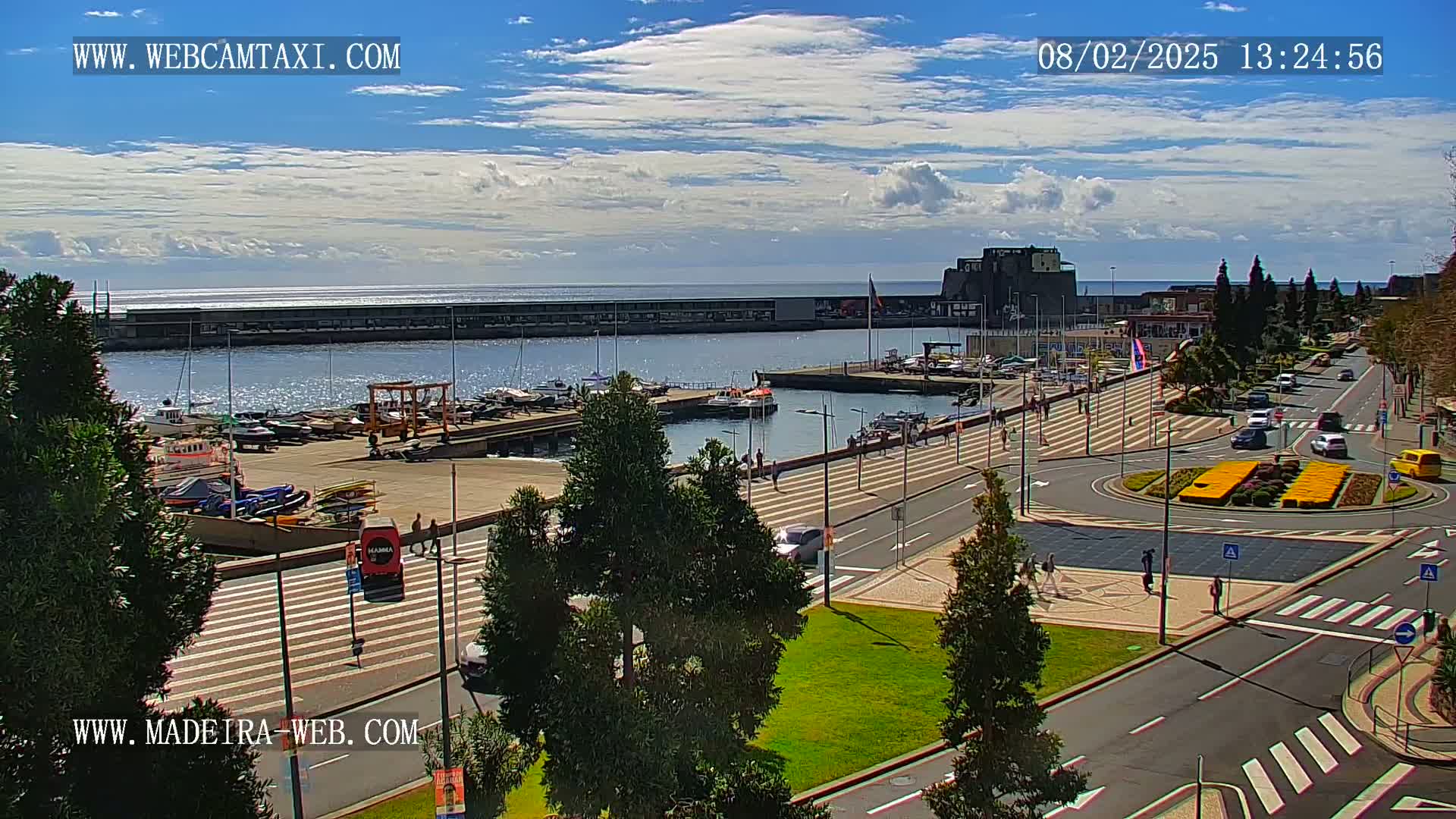 A marina with boats docked, a paved waterfront area with pedestrians and cars, and a bright sunny sky with some clouds.