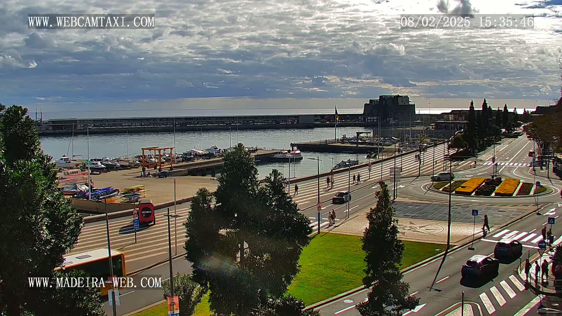 A partly cloudy day overlooking a harbor filled with boats and a paved city area with cars and pedestrians, featuring a roundabout with yellow flowers in the center.