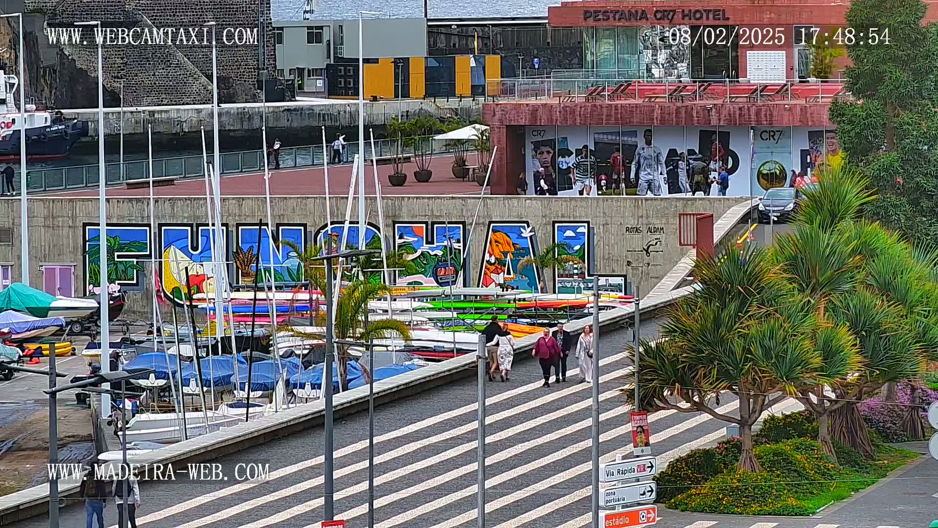 A harbor scene with numerous boats and a large mural of the word "FUNCHAL" on a wall, under partly cloudy skies.