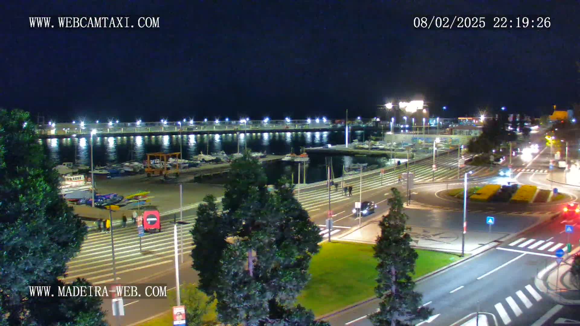 A nighttime, high-angle view of a harbor with boats docked, a surrounding roadway with vehicles, and illuminated landscaping.