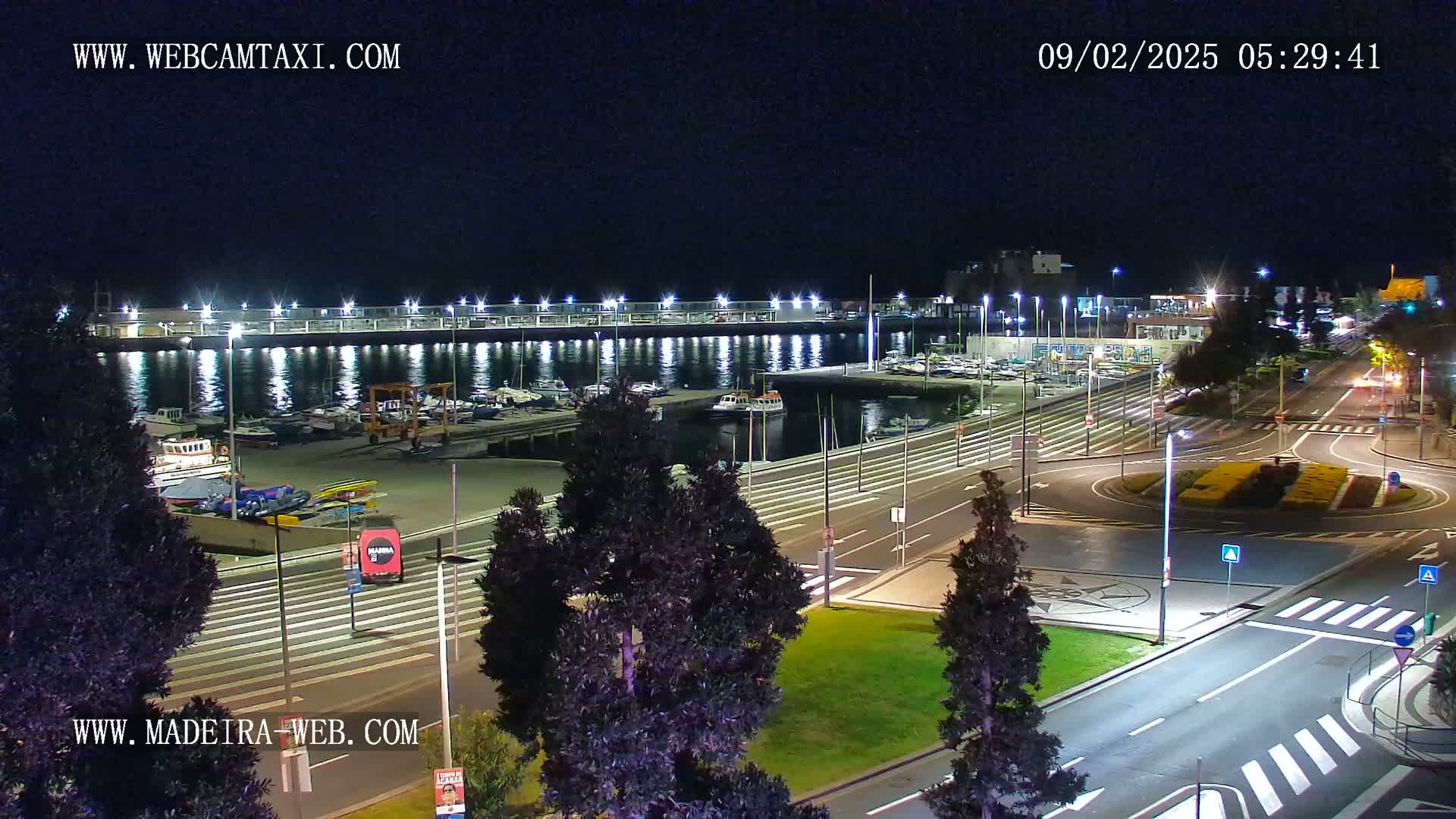 A nighttime view of a harbor filled with boats, adjacent to a roadway with a roundabout, under clear skies.