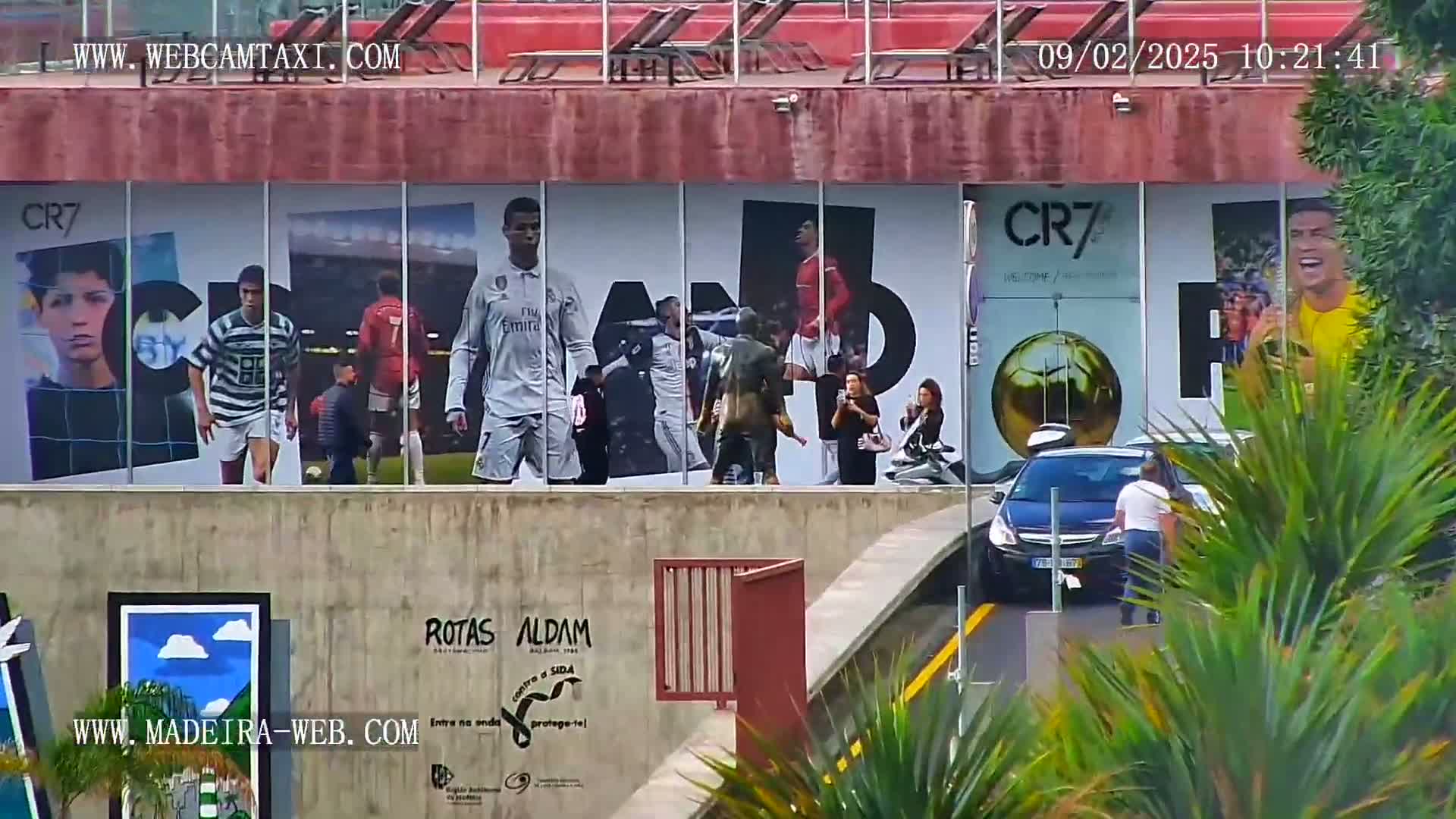 A large building displays a mural of Cristiano Ronaldo throughout his career, and several people are standing outside near parked cars on a sunny day.