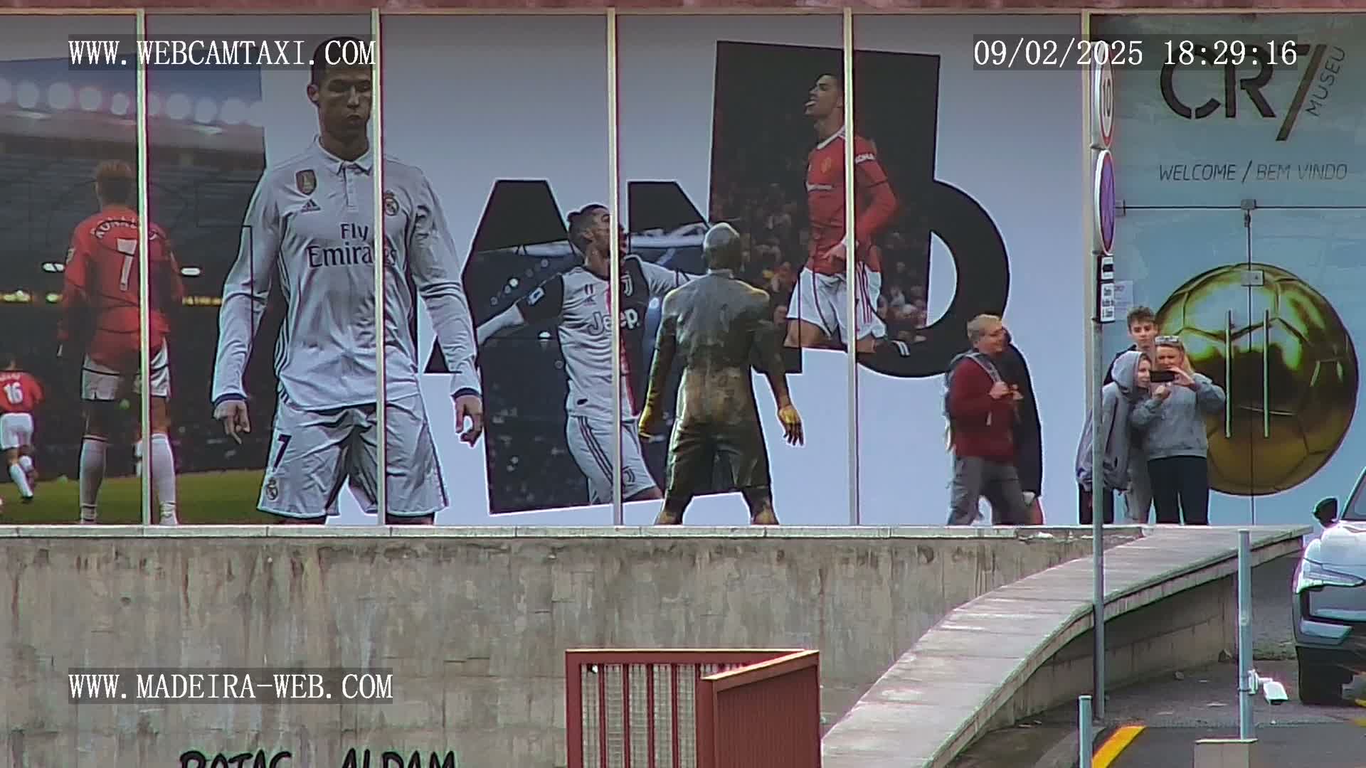 A large building exterior displays images of Cristiano Ronaldo in various soccer jerseys, along with a bronze statue of him; several people stand in front of the building taking photos.