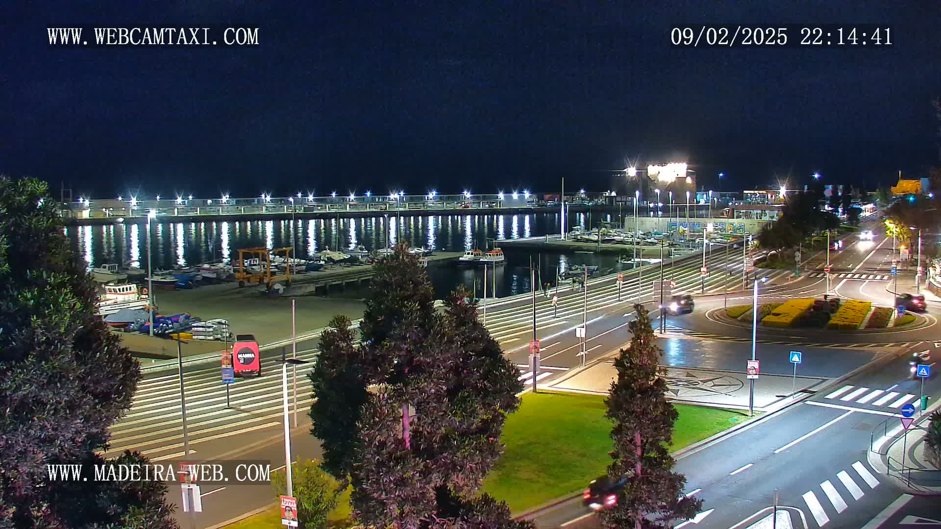 A nighttime view of a harbor filled with boats, next to a roadway with a roundabout, under a clear sky.