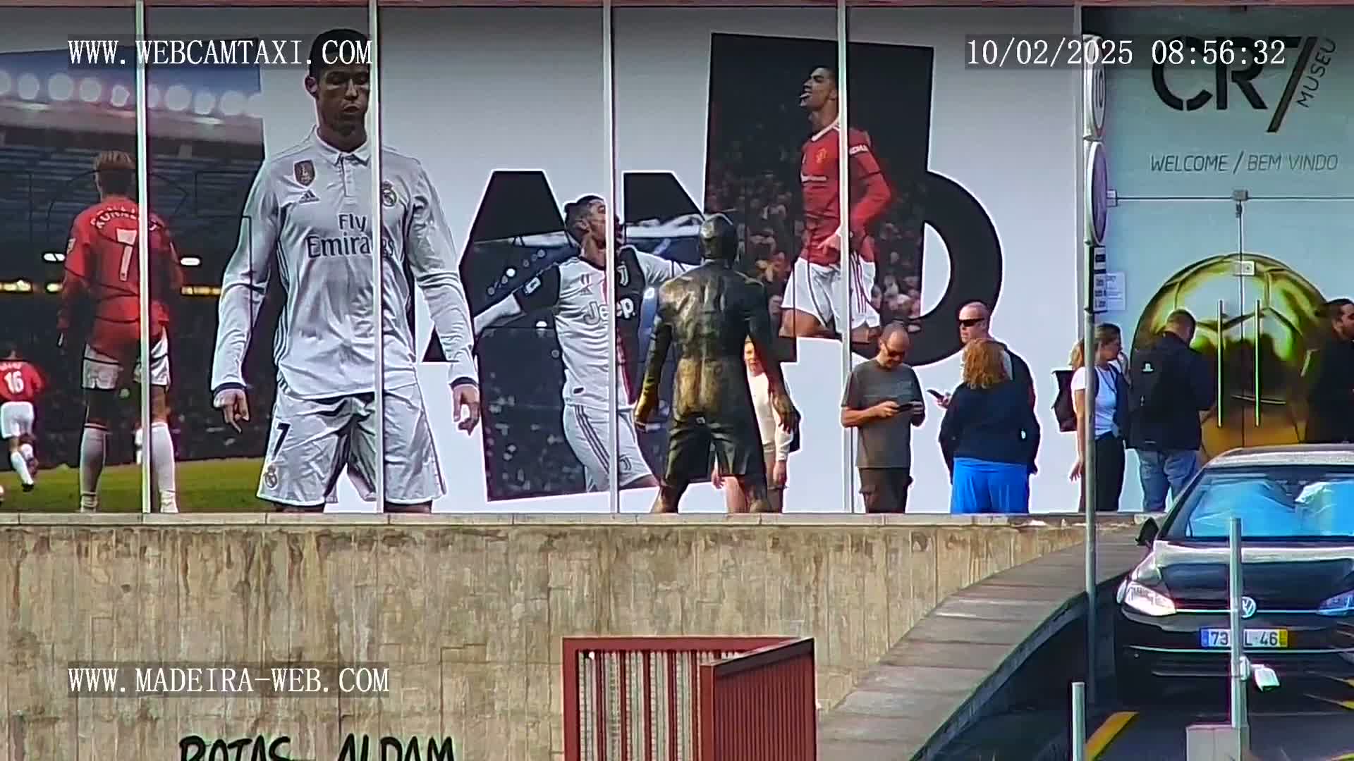 A large outdoor billboard displays images of Cristiano Ronaldo in various soccer uniforms, along with a bronze statue of him, while several people stand in front of it on a sunny day.