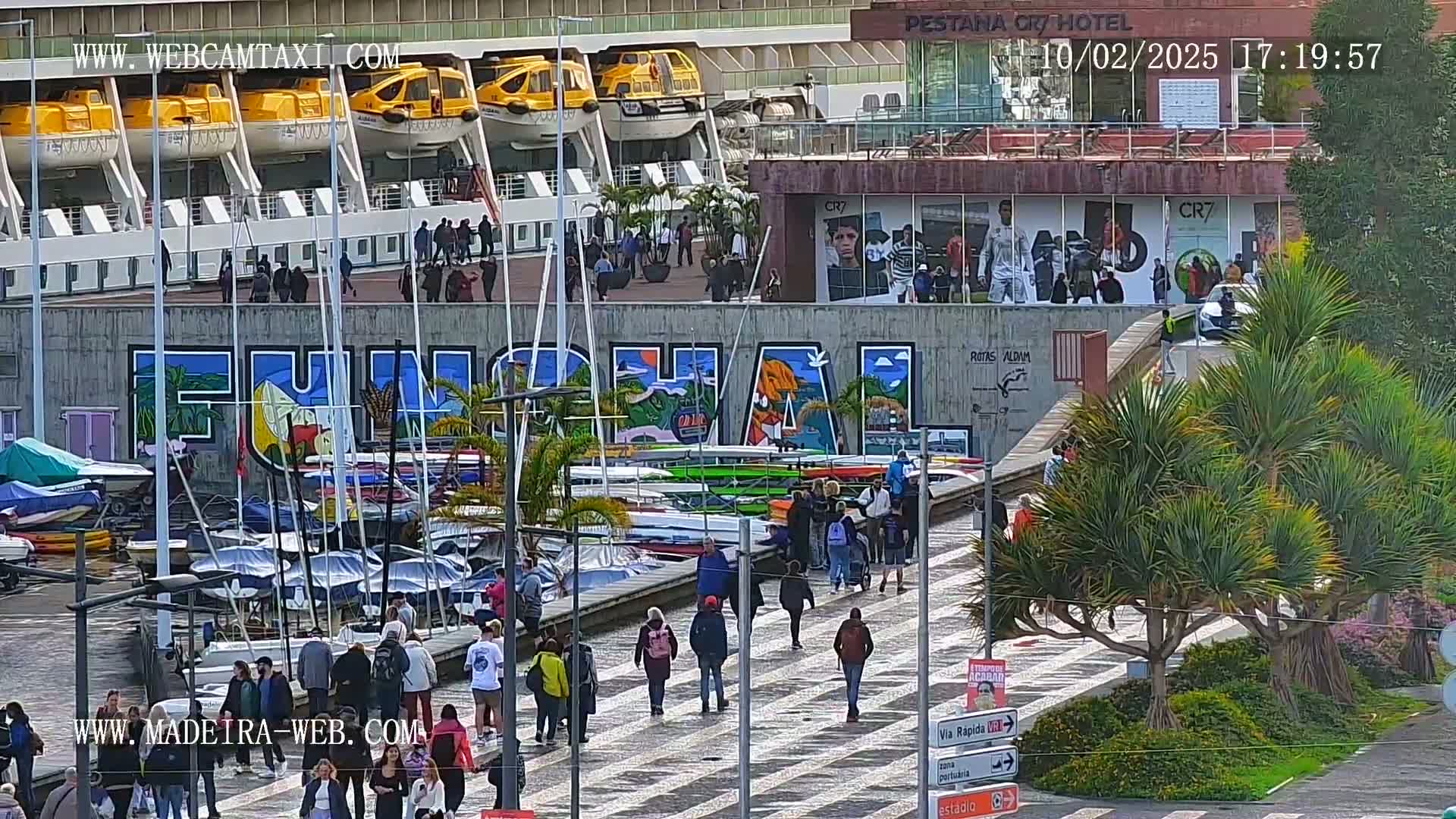 A cruise ship is docked at a harbor with a large mural of the word "FUNCHAL" on a wall, and many people are walking along a paved area near numerous boats and palm trees on a partly cloudy day.