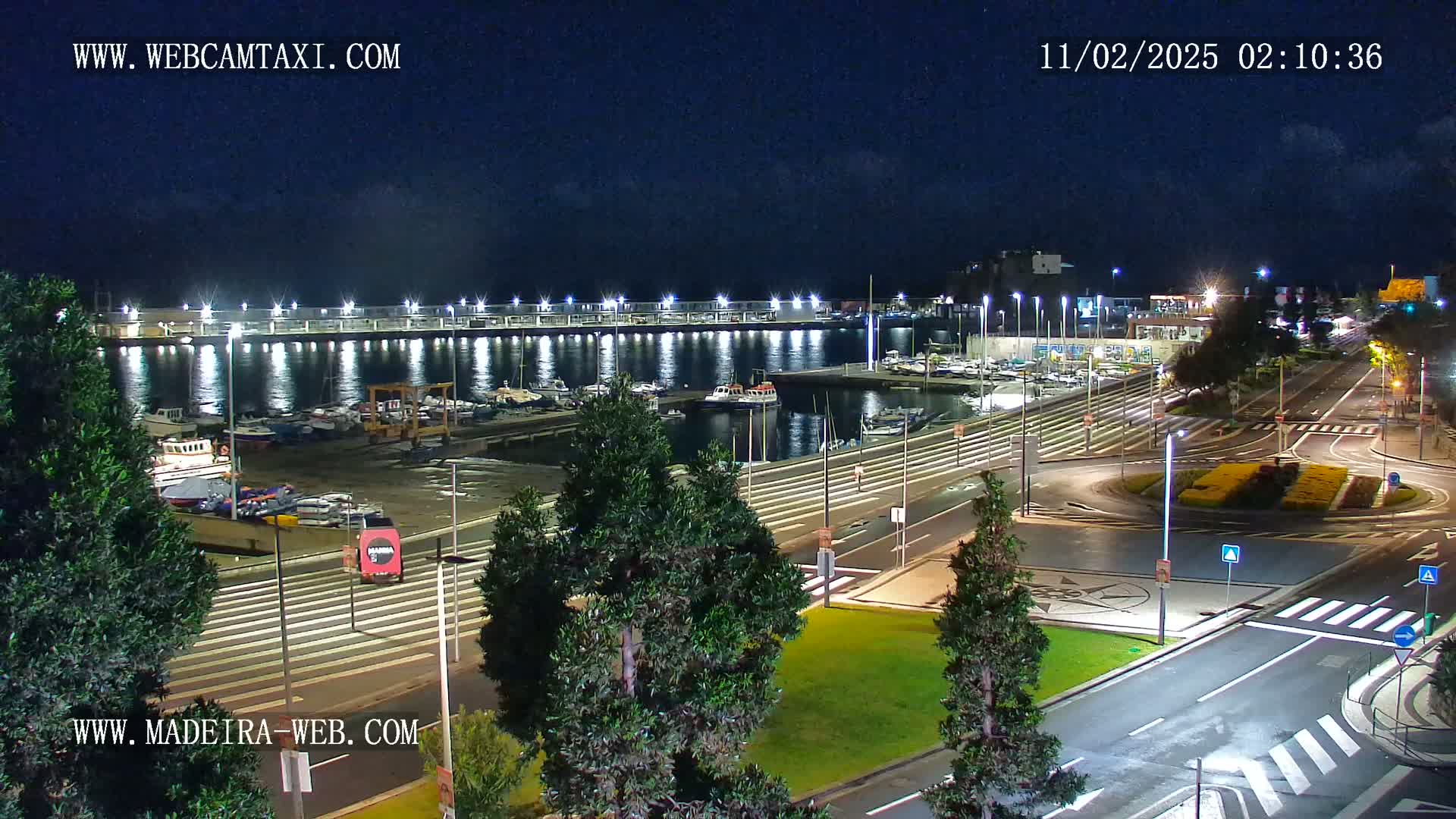 A nighttime view of a harbor filled with boats, adjacent to a well-lit roadway and roundabout, under a mostly clear sky.