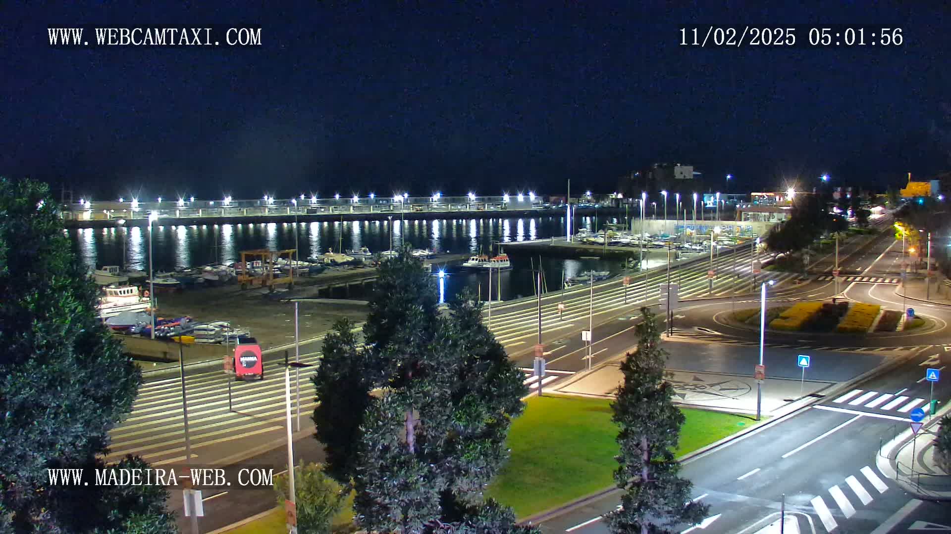 A nighttime view of a harbor filled with boats, next to a roadway with a traffic circle, under a clear, dark sky.