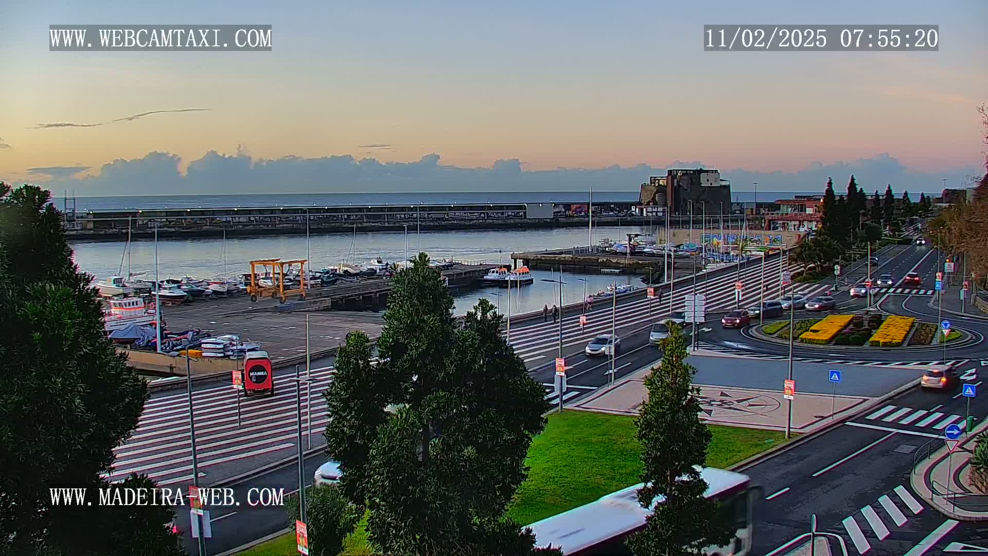 A harbor with boats docked, a roadway with cars, and a roundabout are visible under a partly cloudy, dusky sky.