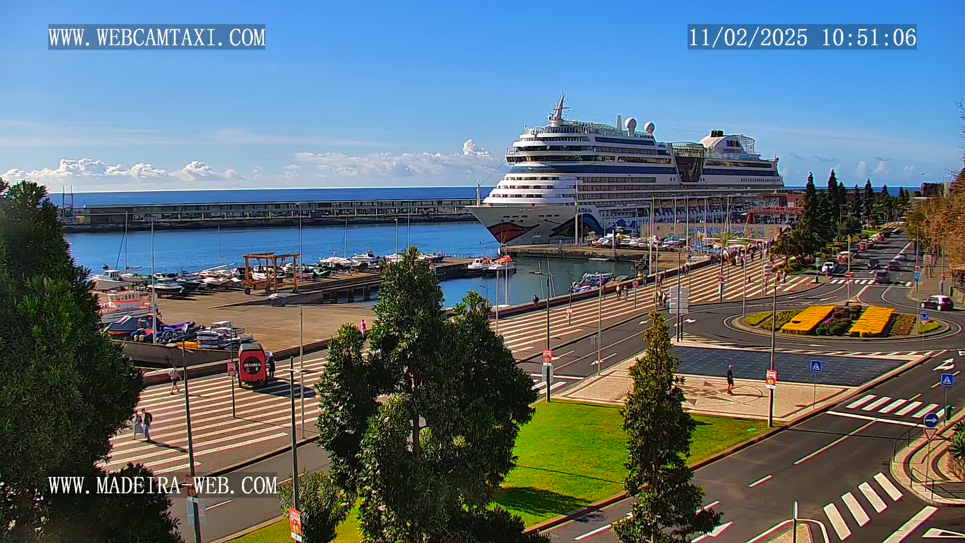 A large cruise ship is docked at a harbor on a sunny day, with smaller boats moored nearby and pedestrians walking along a paved area.