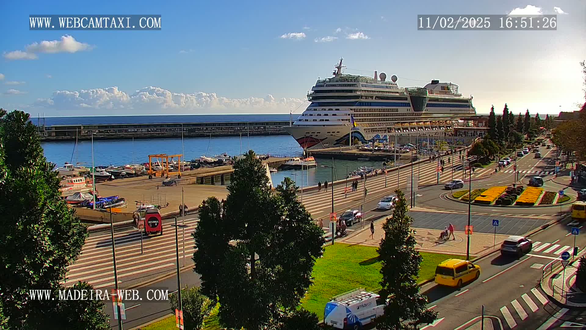 A large cruise ship is docked at a harbor on a sunny day with some clouds in the sky, and there are cars and people on the streets nearby.