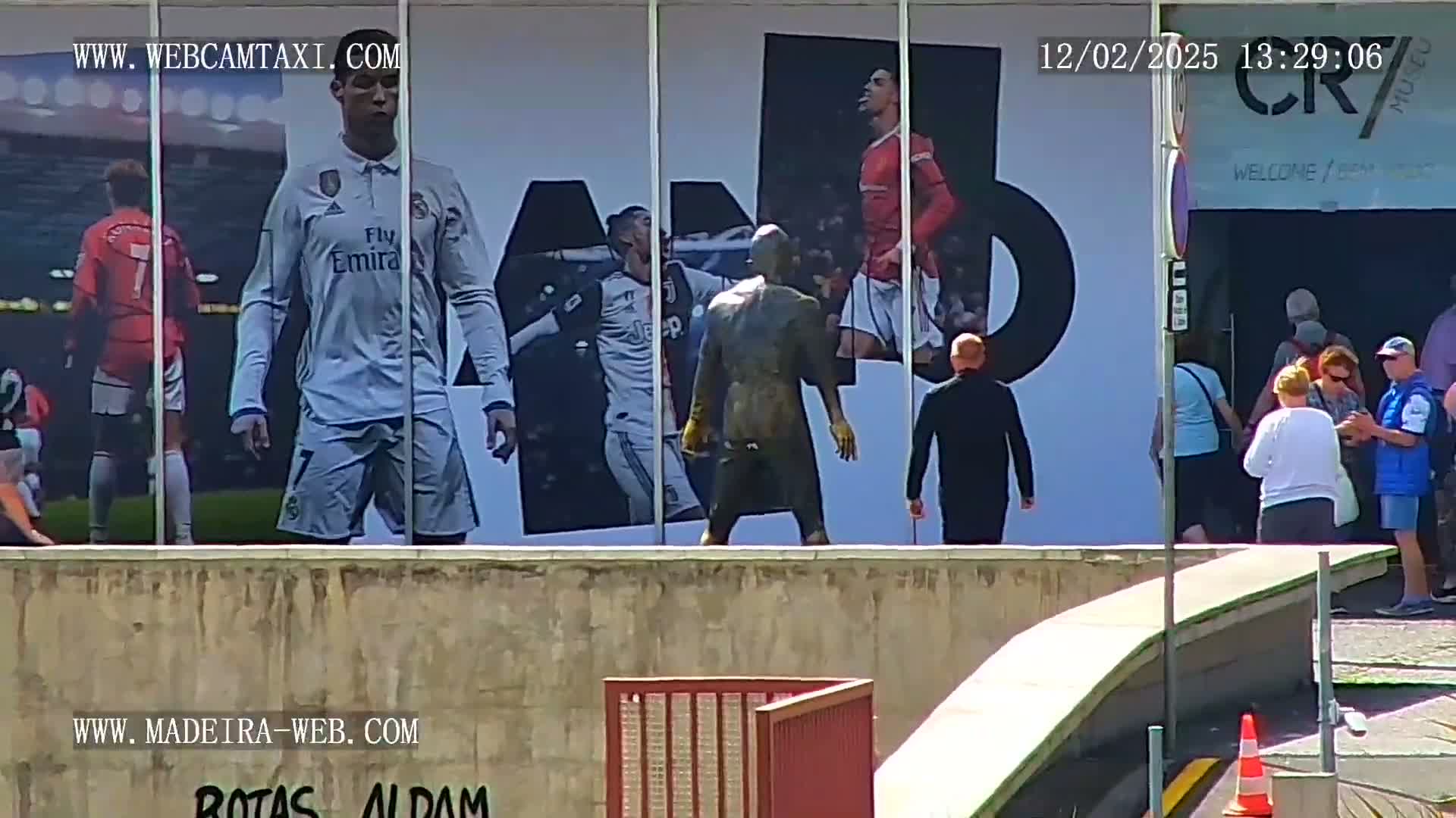 A large outdoor mural depicting several soccer players is viewed from across a low wall, with several people standing in front of the mural on a sunny day.