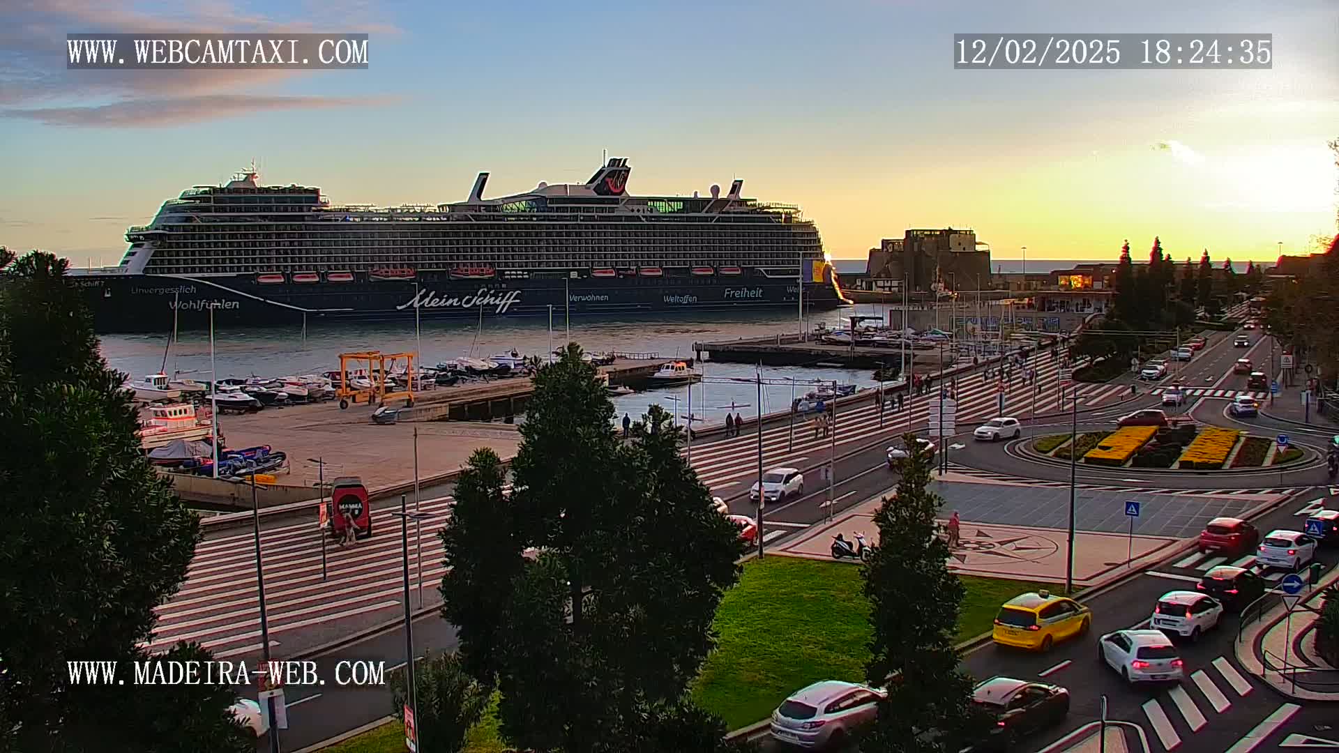 A large cruise ship is docked at a harbor during sunset, with several cars driving on a roadway next to the water.