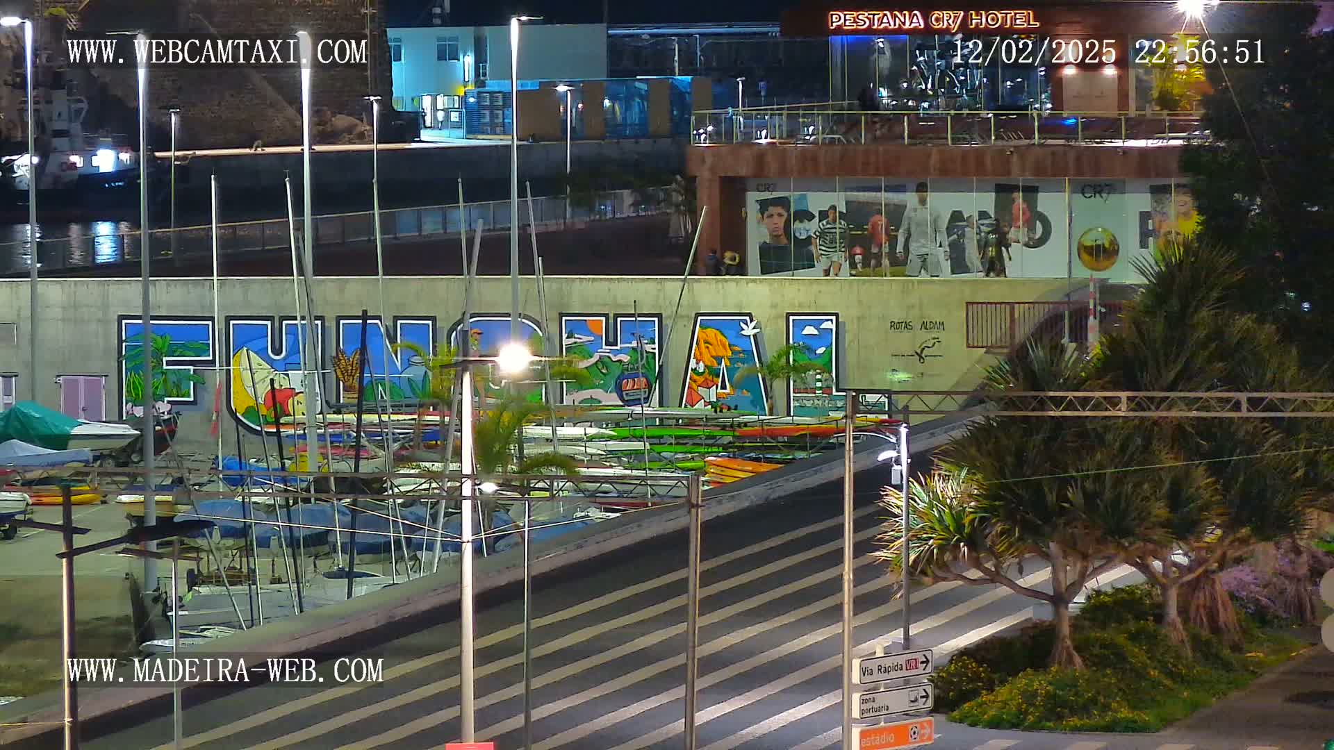 A nighttime view of a marina with numerous boats and kayaks docked, featuring a large mural of the word "Funchal," situated beneath a building with illuminated advertisements.