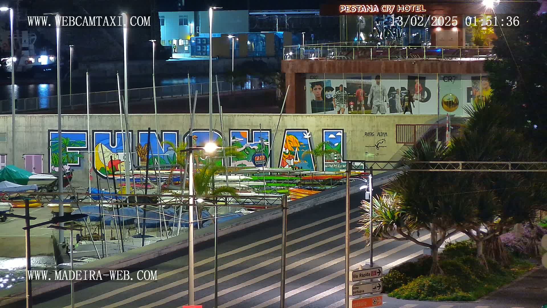 A nighttime view of a harbor filled with boats, a large mural reading "Funchal," and palm trees lining a roadway.