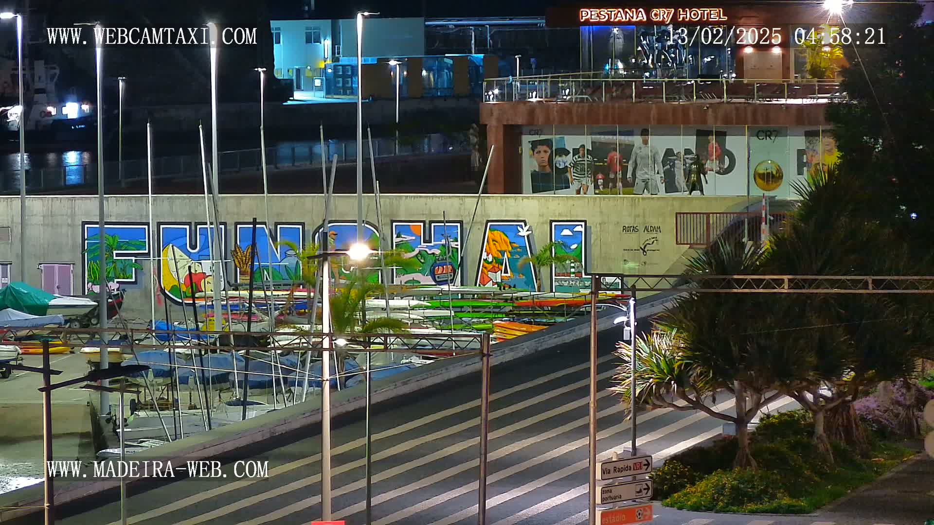 A nighttime view of a harbor with numerous boats docked alongside a wall featuring large graffiti art of the word "FUNCHAL."