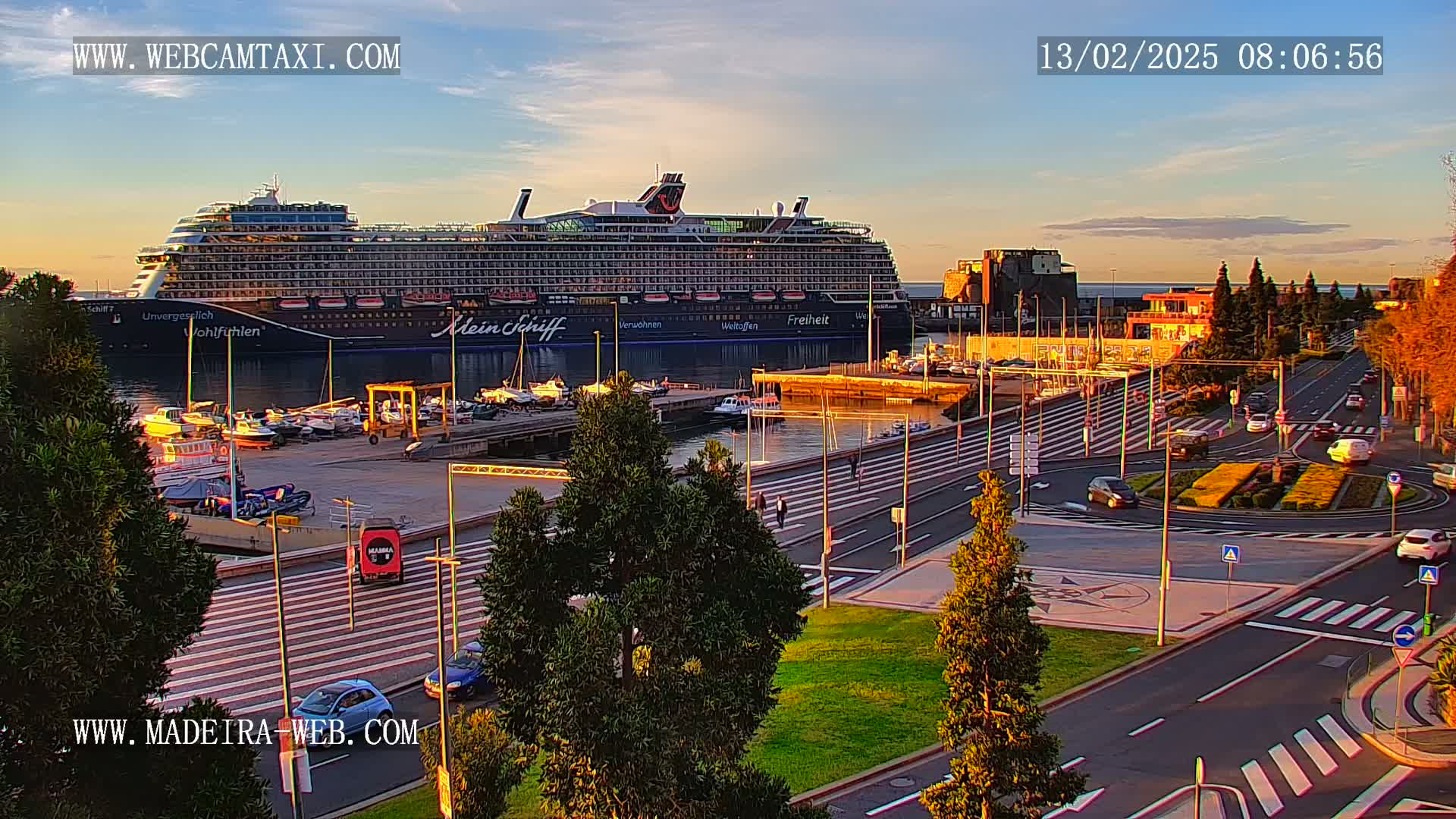 A large cruise ship is docked at a harbor in the evening, with cars driving on a nearby road under a clear, sunset sky.