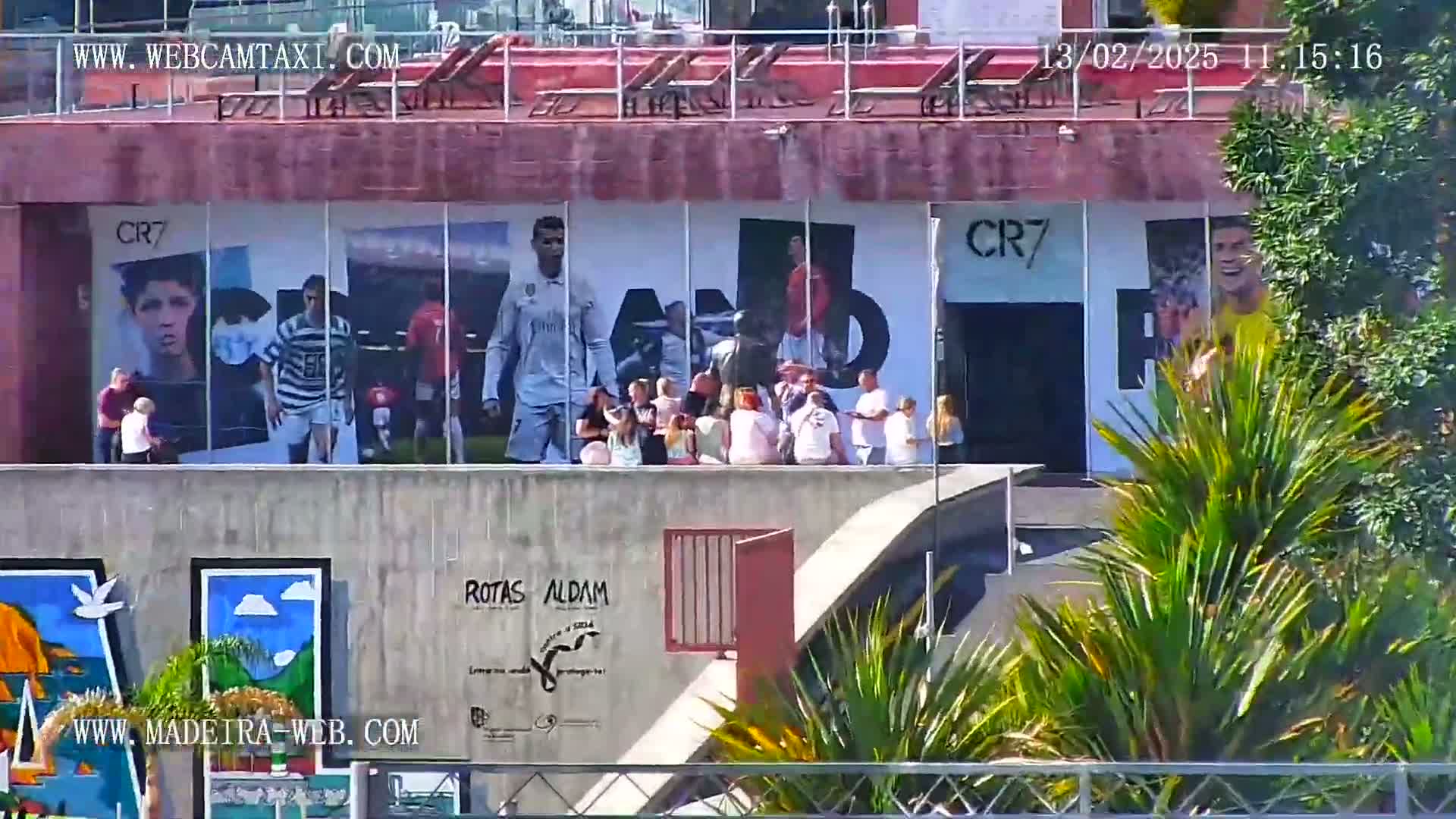 A group of people stand on a rooftop overlooking a building adorned with large murals of Cristiano Ronaldo and other soccer players, on a sunny day.