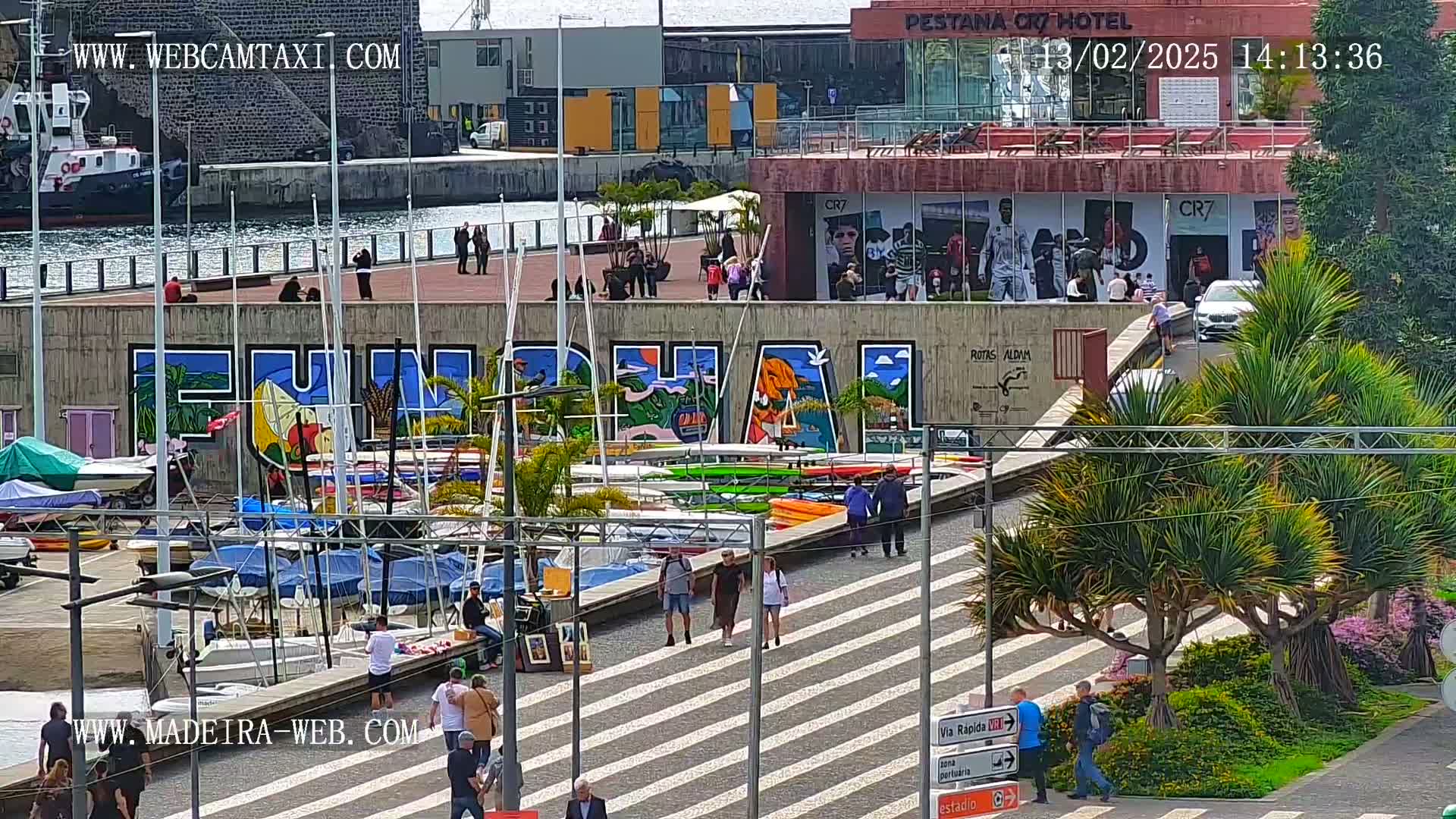 A waterfront scene with a large mural of "Funchal" on a wall overlooking a harbor filled with sailboats and kayaks, under a sunny sky.